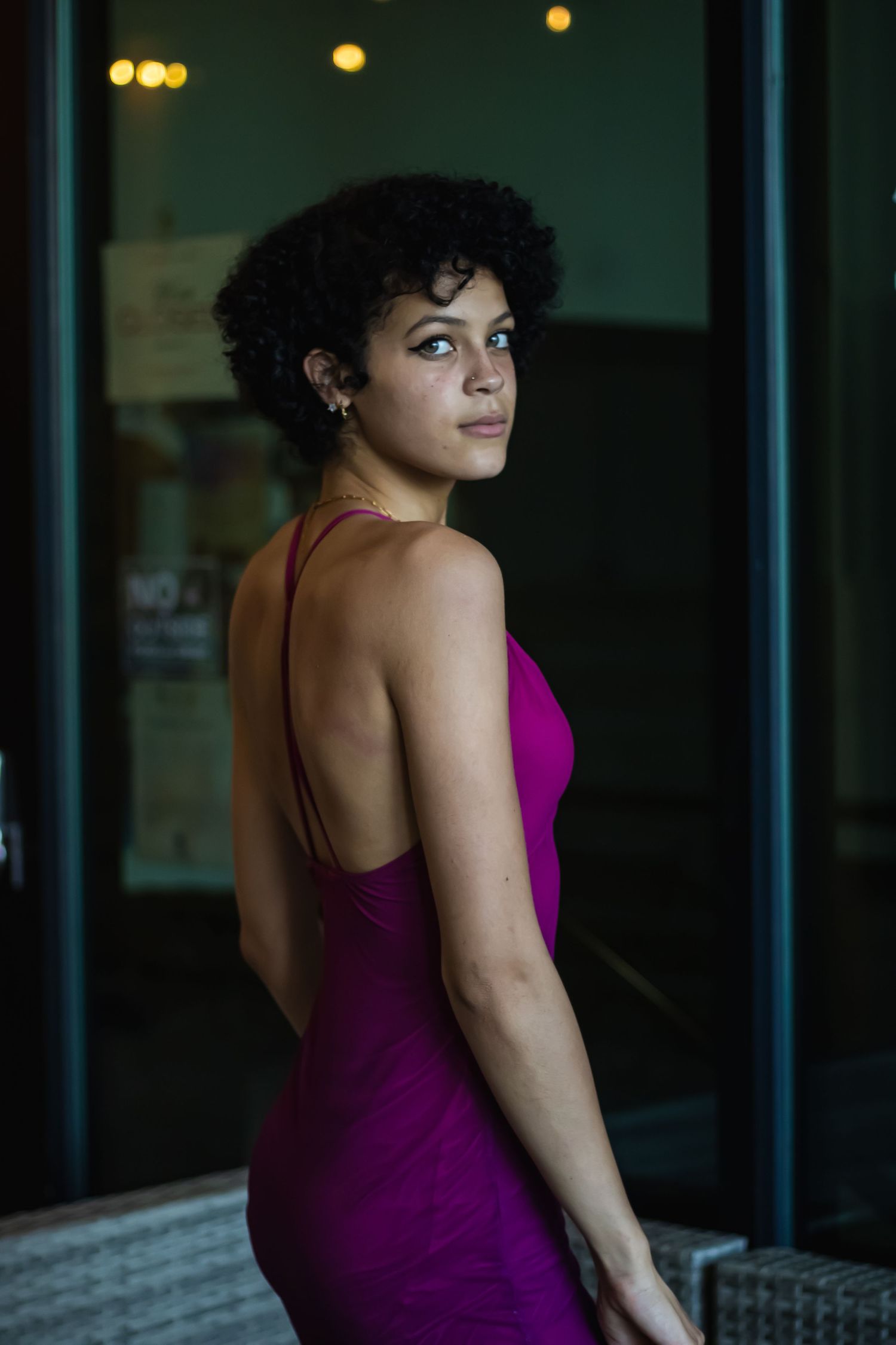 A young woman with short curly hair poses in a backless magenta dress, glancing over her shoulder near a glass door.