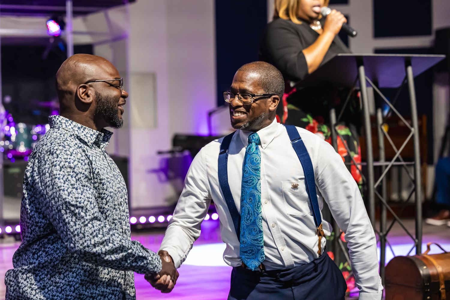 Two well-dressed men share a warm handshake and smile at an indoor celebration event.