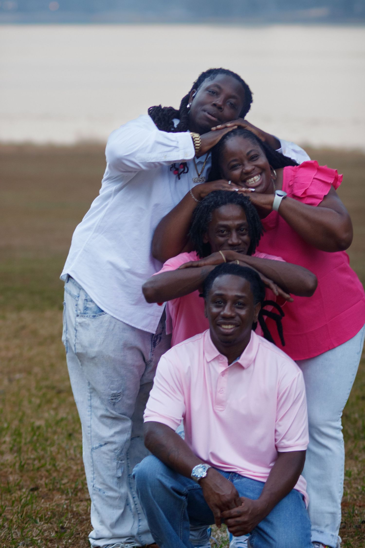 Four joyful family members pose playfully outdoors near a lake, stacking heads in a fun portrait on a grassy field.