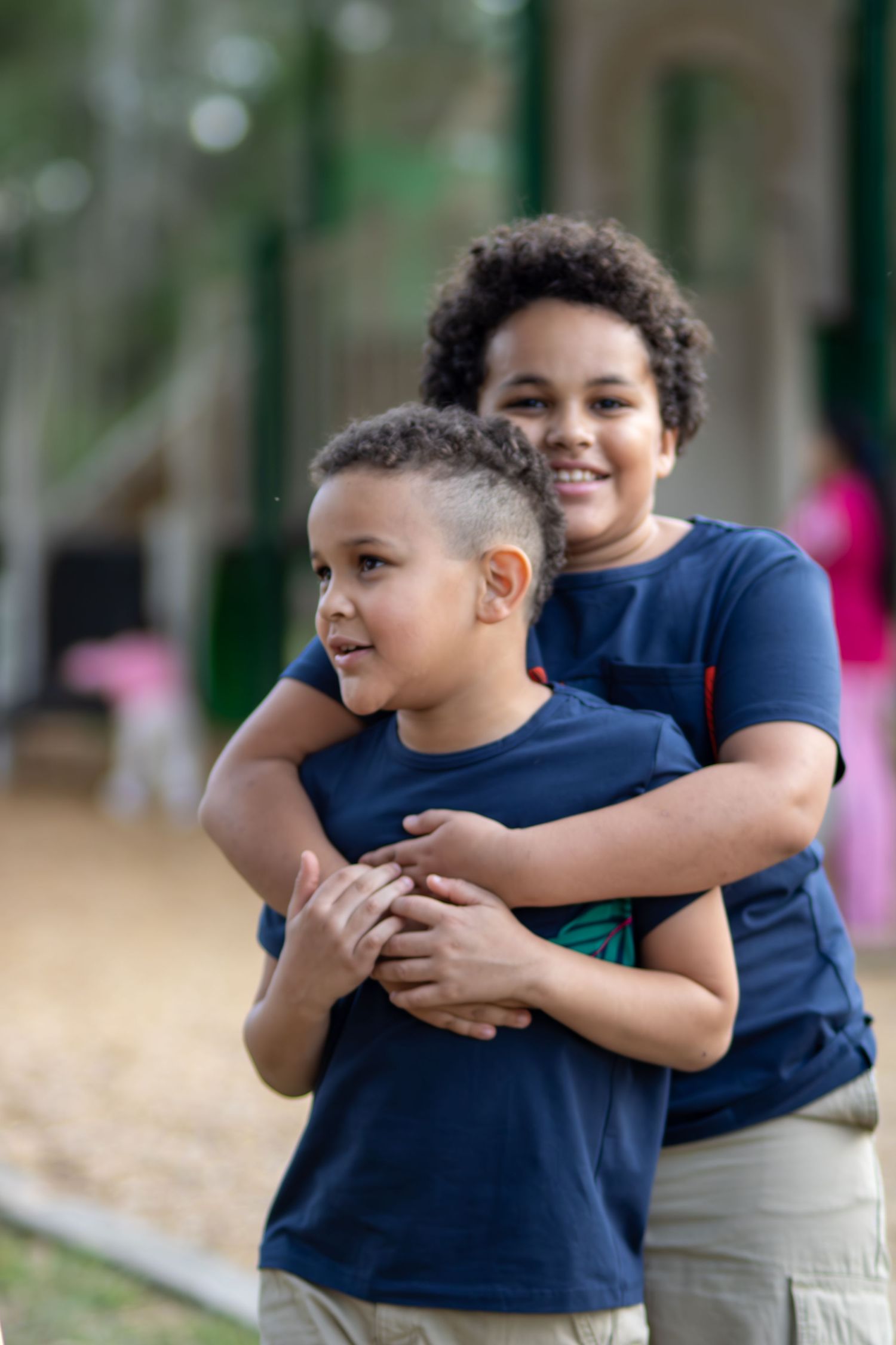 Two young boys in matching navy blue shirts share a warm hug outdoors on a playground.
