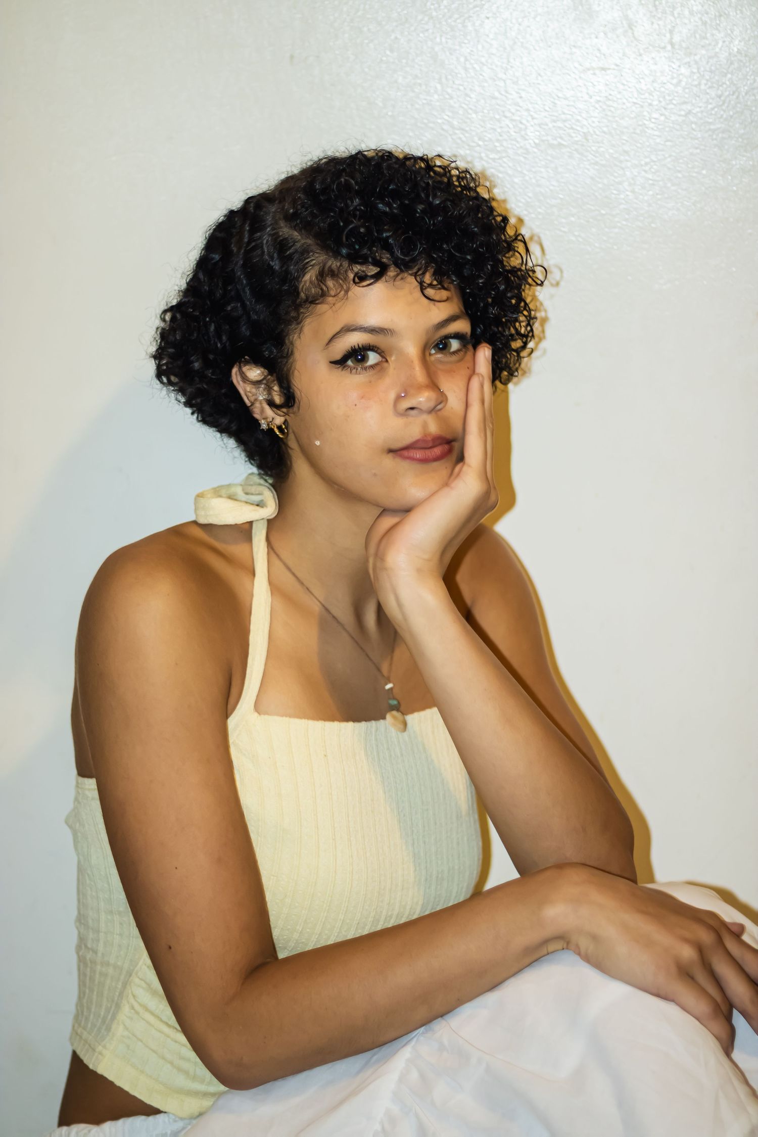 Young woman with curly hair in a yellow crop top resting her chin on her hand against a white wall.