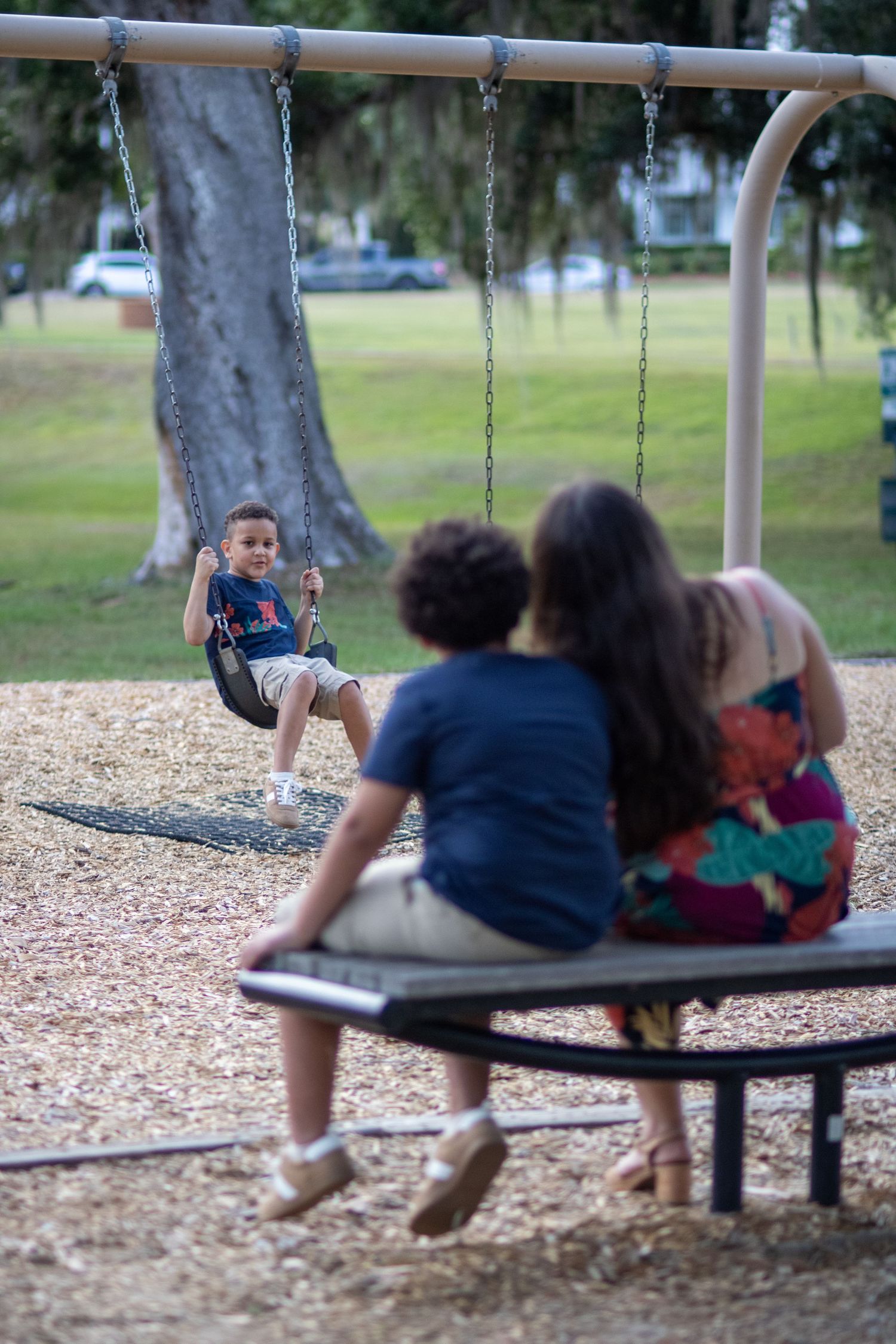 A young boy swings happily at a park playground while two family members watch from a nearby bench.