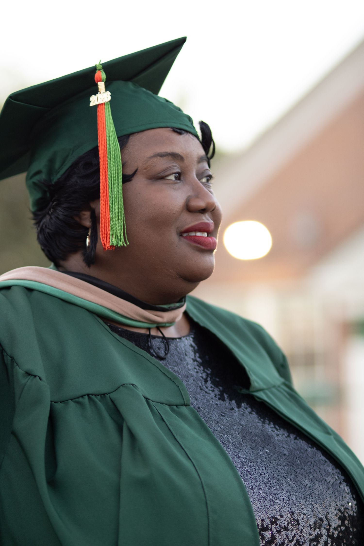 Smiling woman in green graduation cap and gown celebrating her academic achievement outdoors.