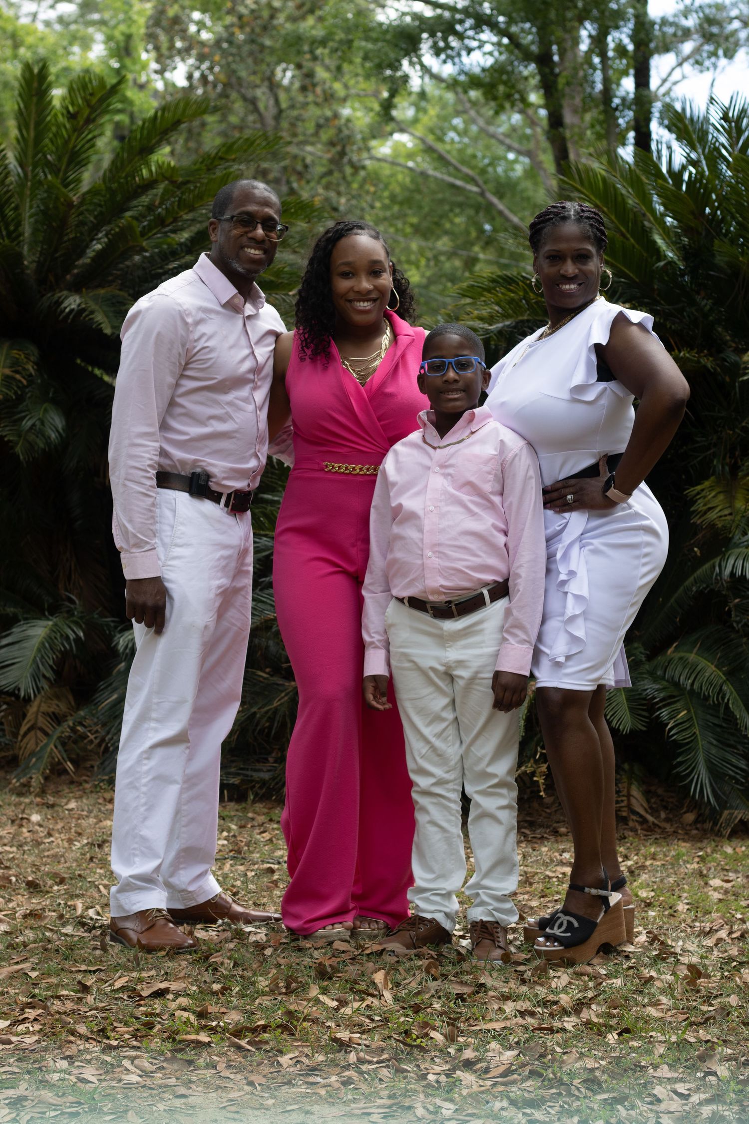 A family of four dressed in pink and white poses together outdoors among lush greenery.