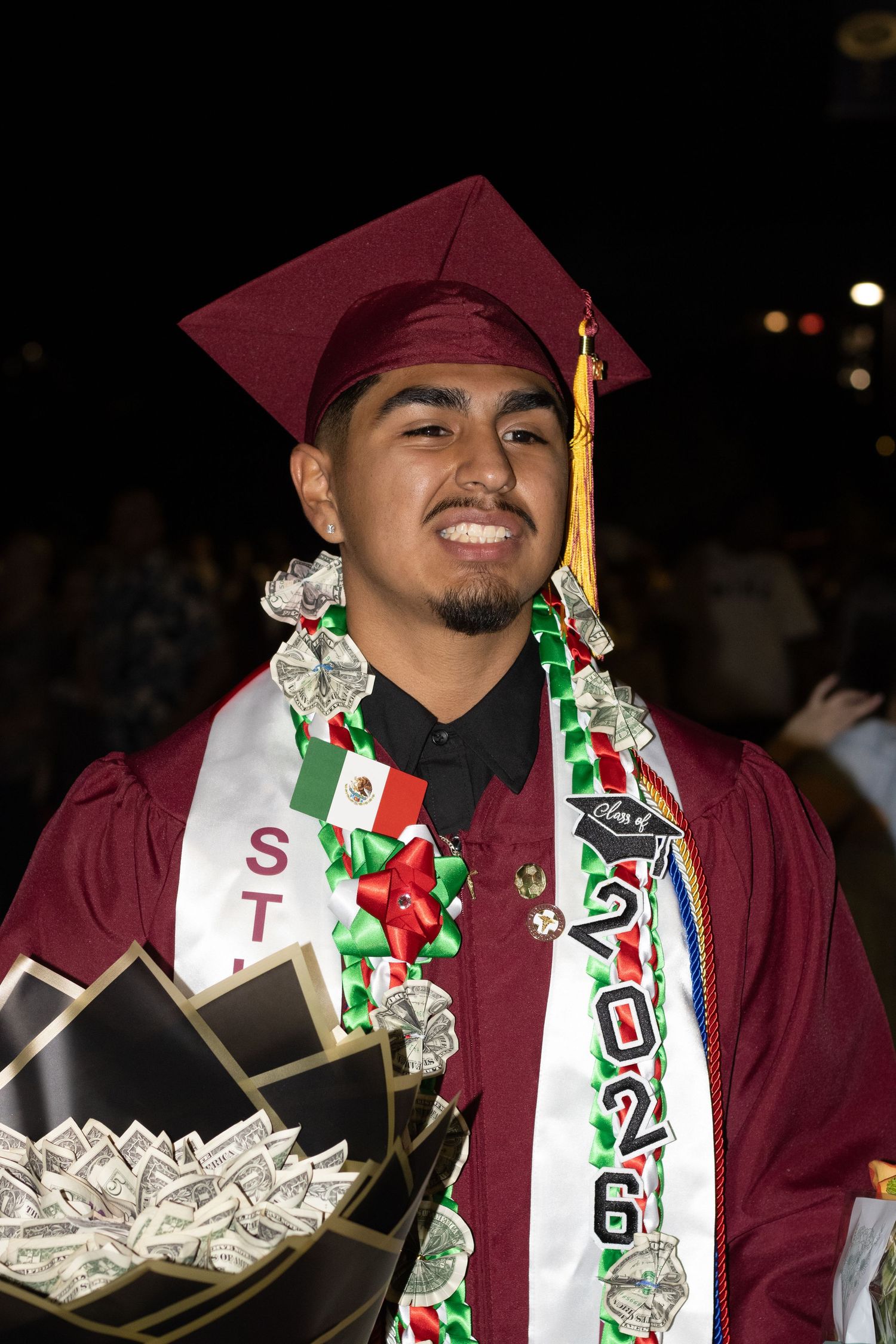 A smiling graduate in maroon cap and gown holds a money bouquet with festive sashes.