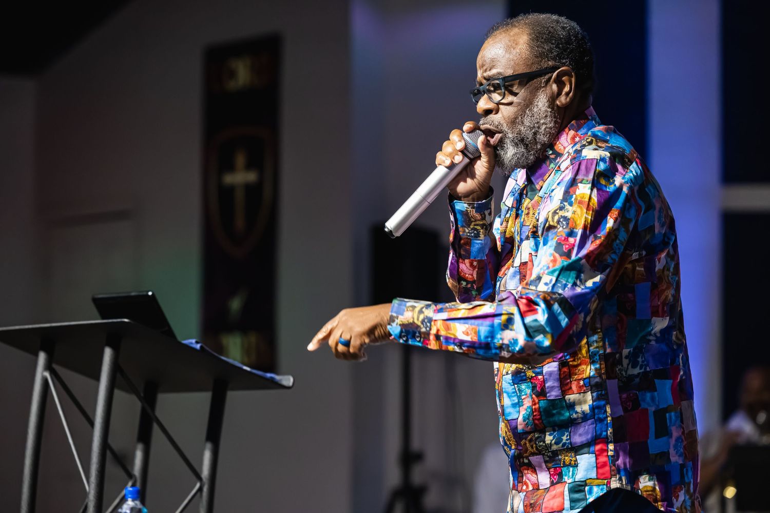 A man in a colorful patchwork shirt speaks passionately into a microphone on stage.