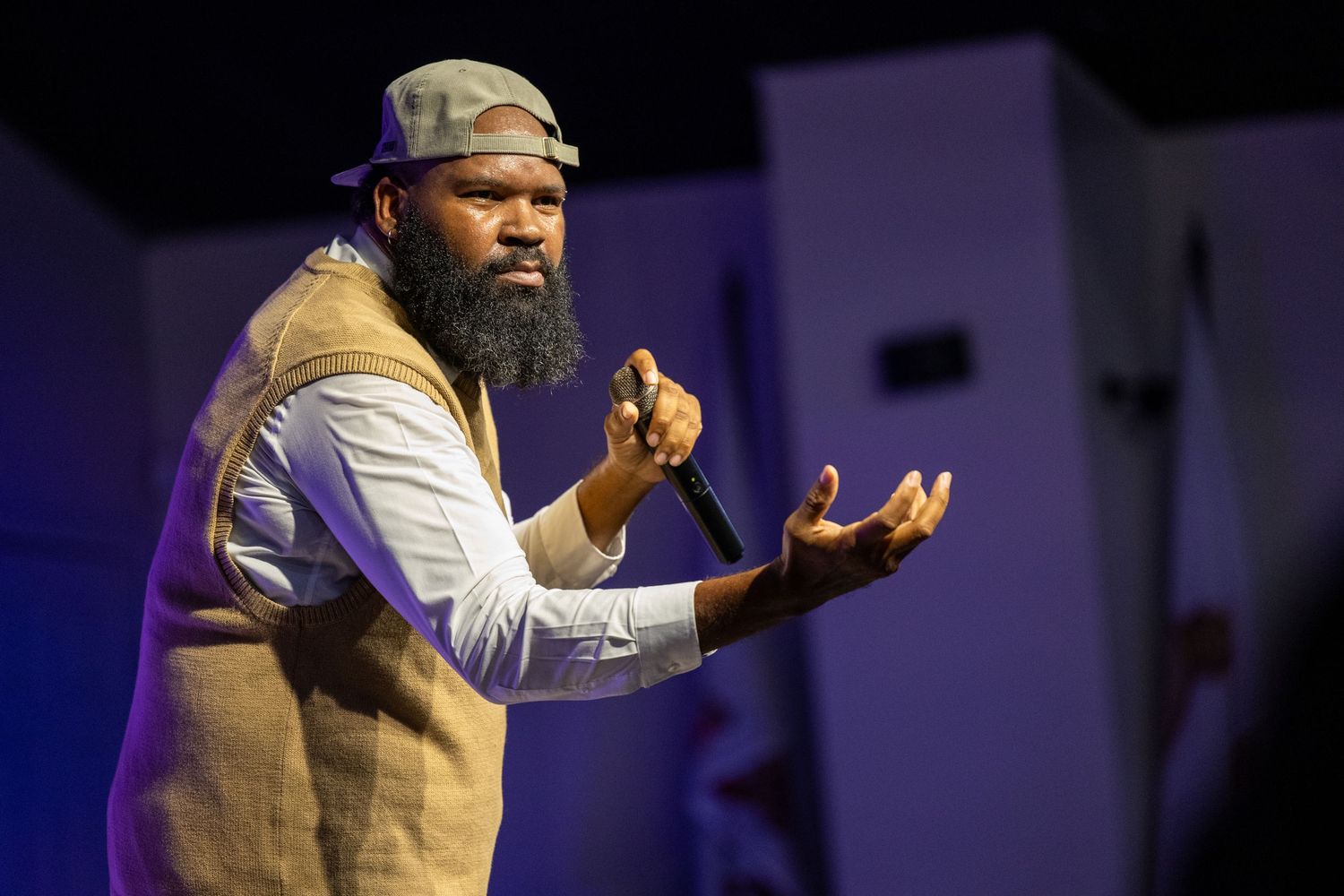Bearded Black man in gray hat and vest speaks passionately into microphone on stage under blue lights.