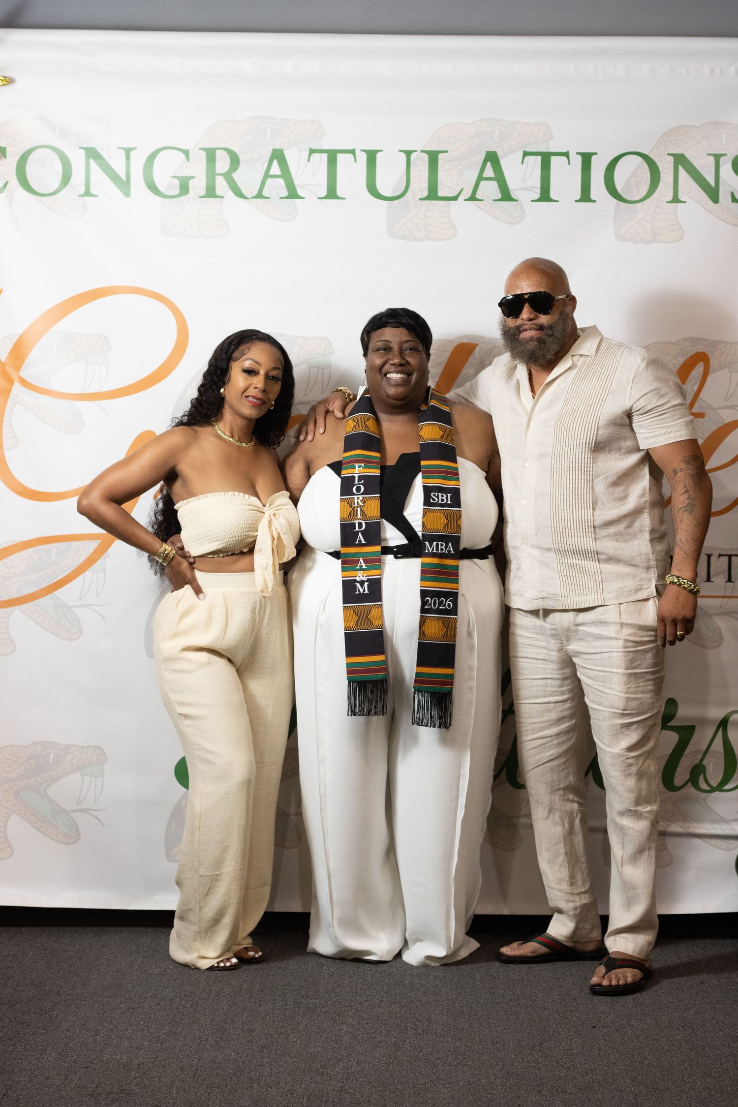 Three people pose together in front of a Congratulations banner at a graduation party.
