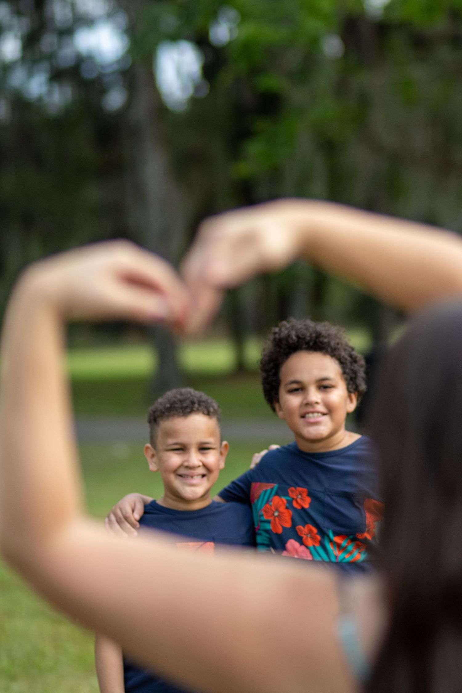 Two smiling boys in navy shirts posed in a park, framed by a heart shape made with someone's hands in the foreground.