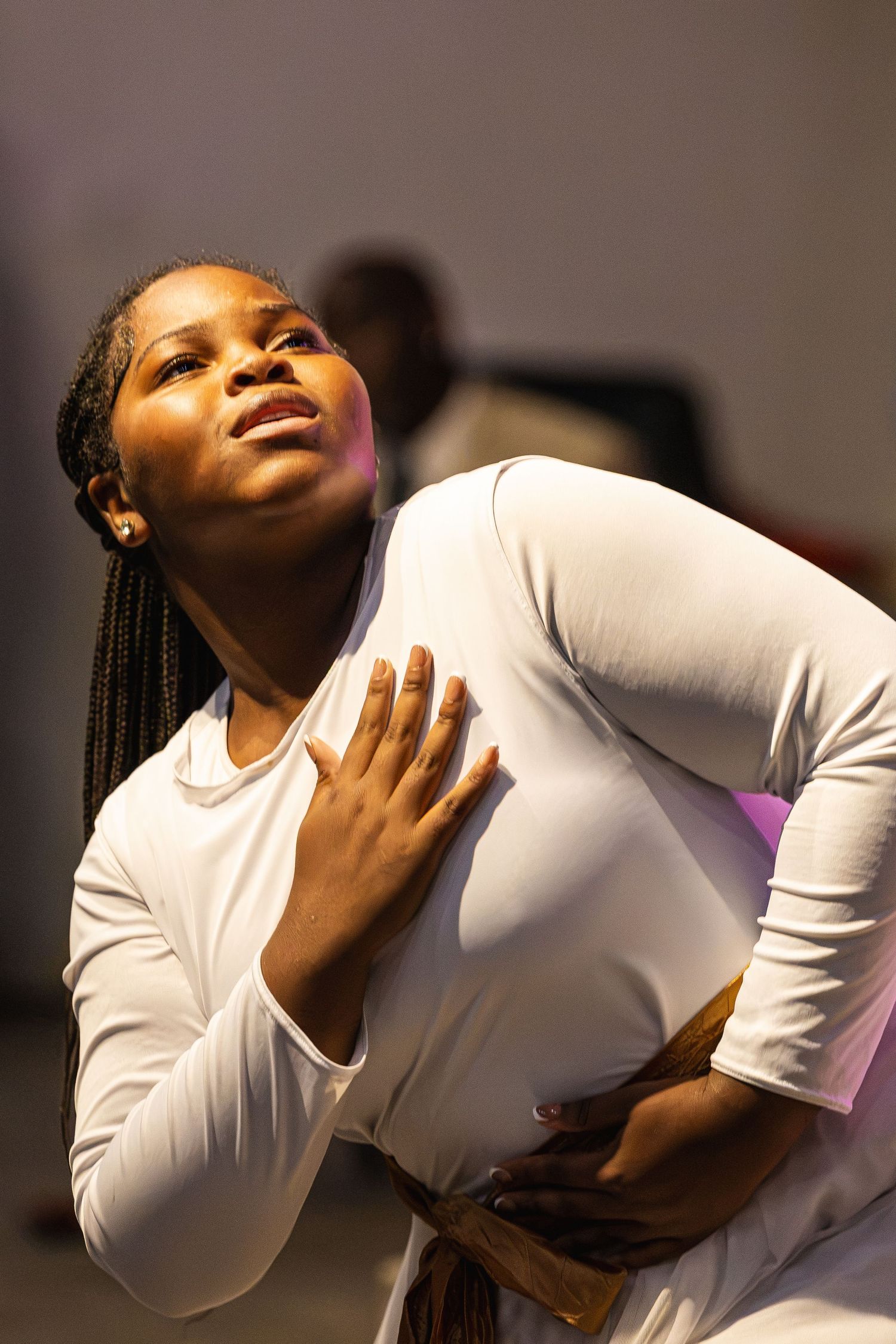Young Black woman in white long-sleeve top performs expressive dance, hand on chest, looking upward with emotion.