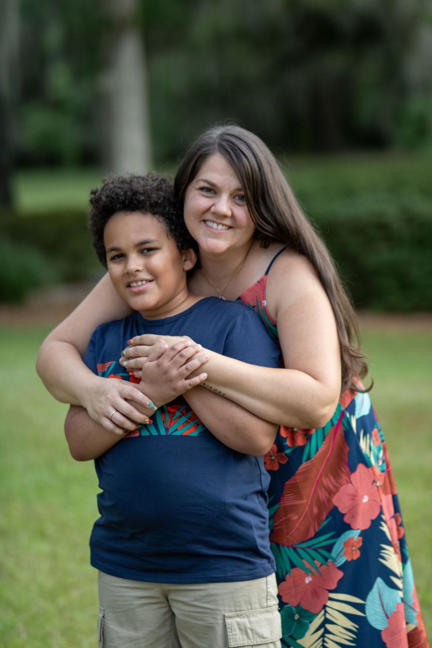 A smiling woman in a floral dress hugs a young boy in a navy shirt outdoors in a lush green park.