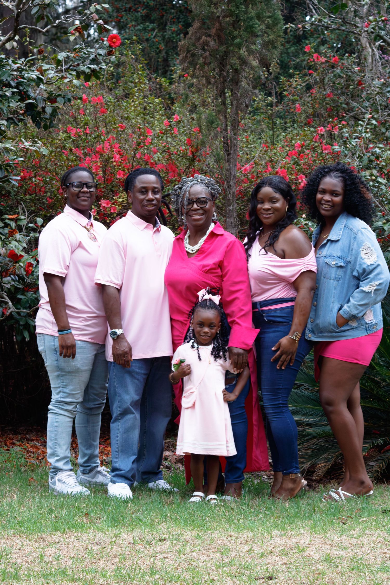 Family of six posing outdoors in coordinating pink outfits with red flowering bushes in the background.