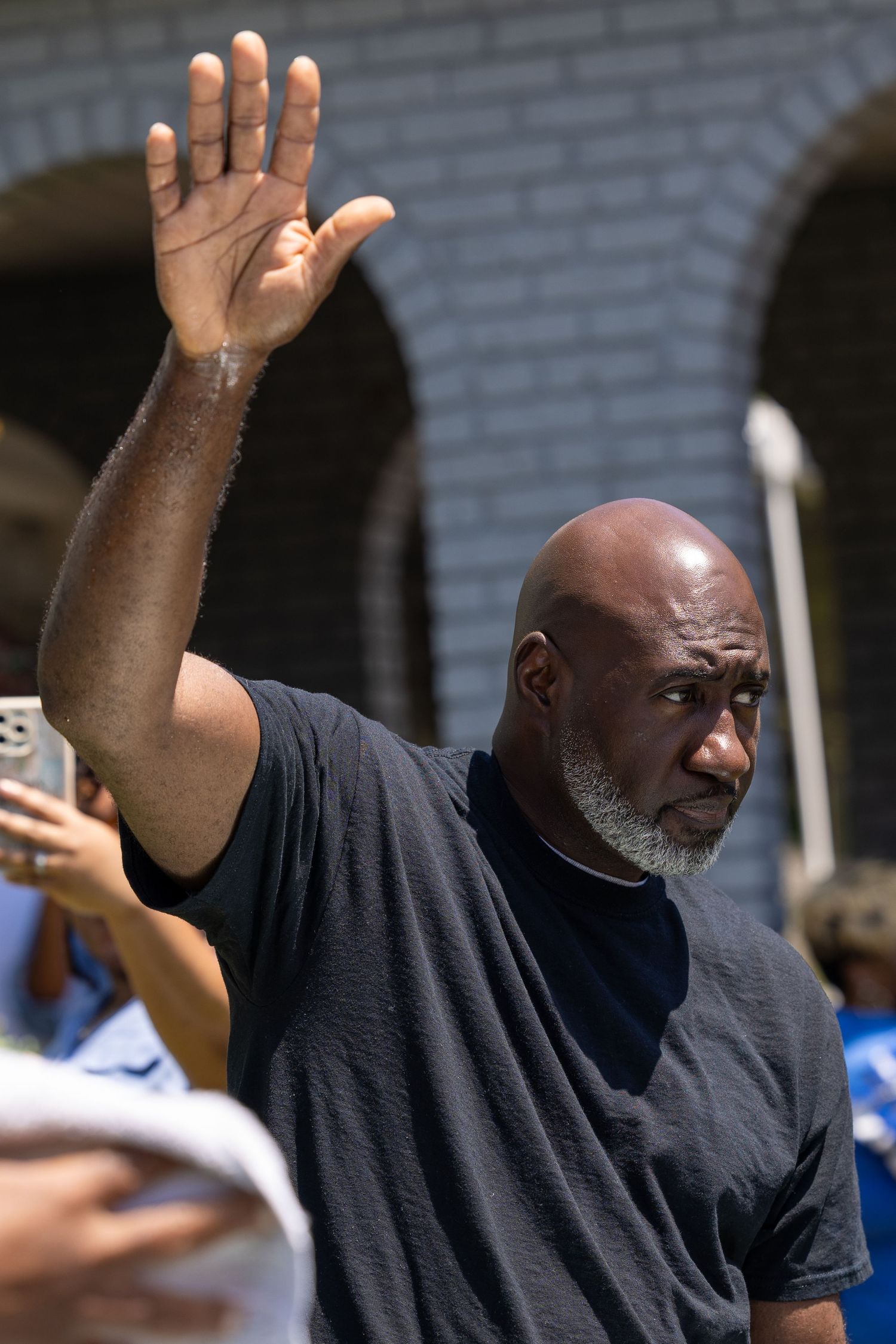 Bald man with gray beard raises hand while standing outdoors in a crowd on a sunny day.