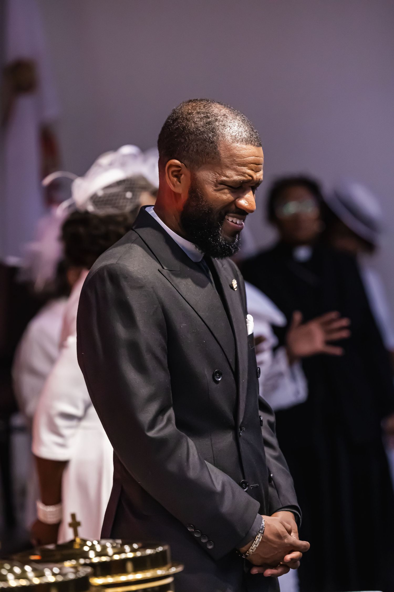 A well-dressed man in a dark suit smiles warmly during a formal church ceremony with congregants in background.