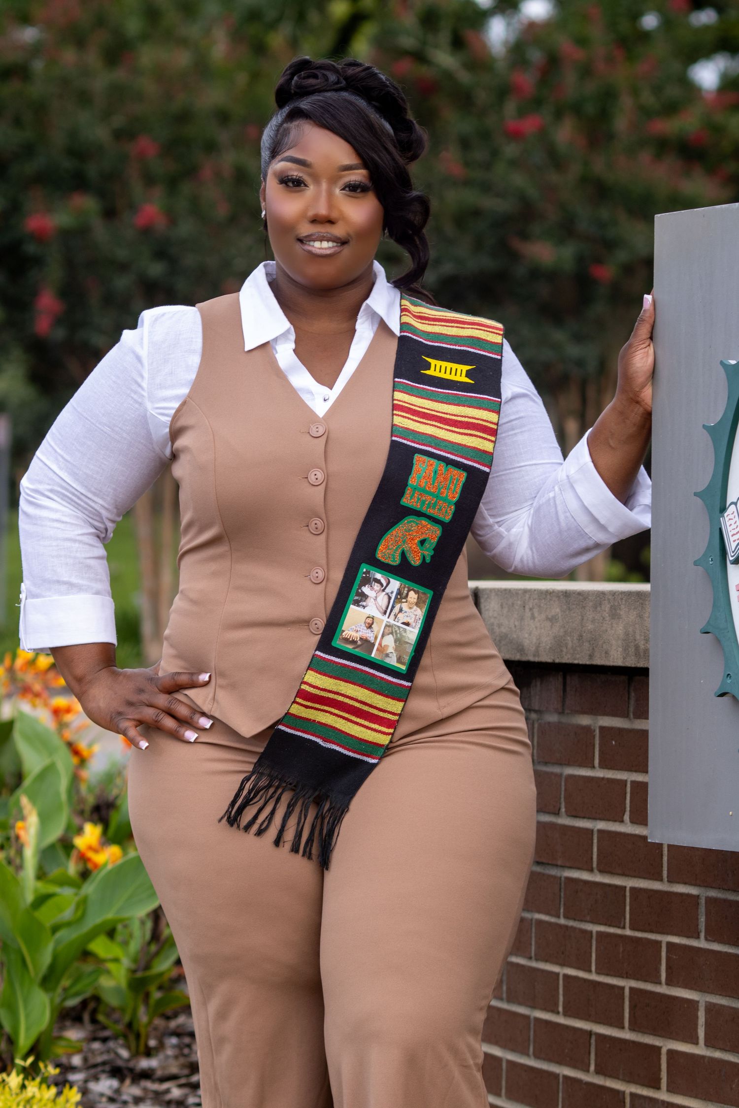 A woman in a tan vest suit wearing a colorful kente graduation stole poses outdoors.