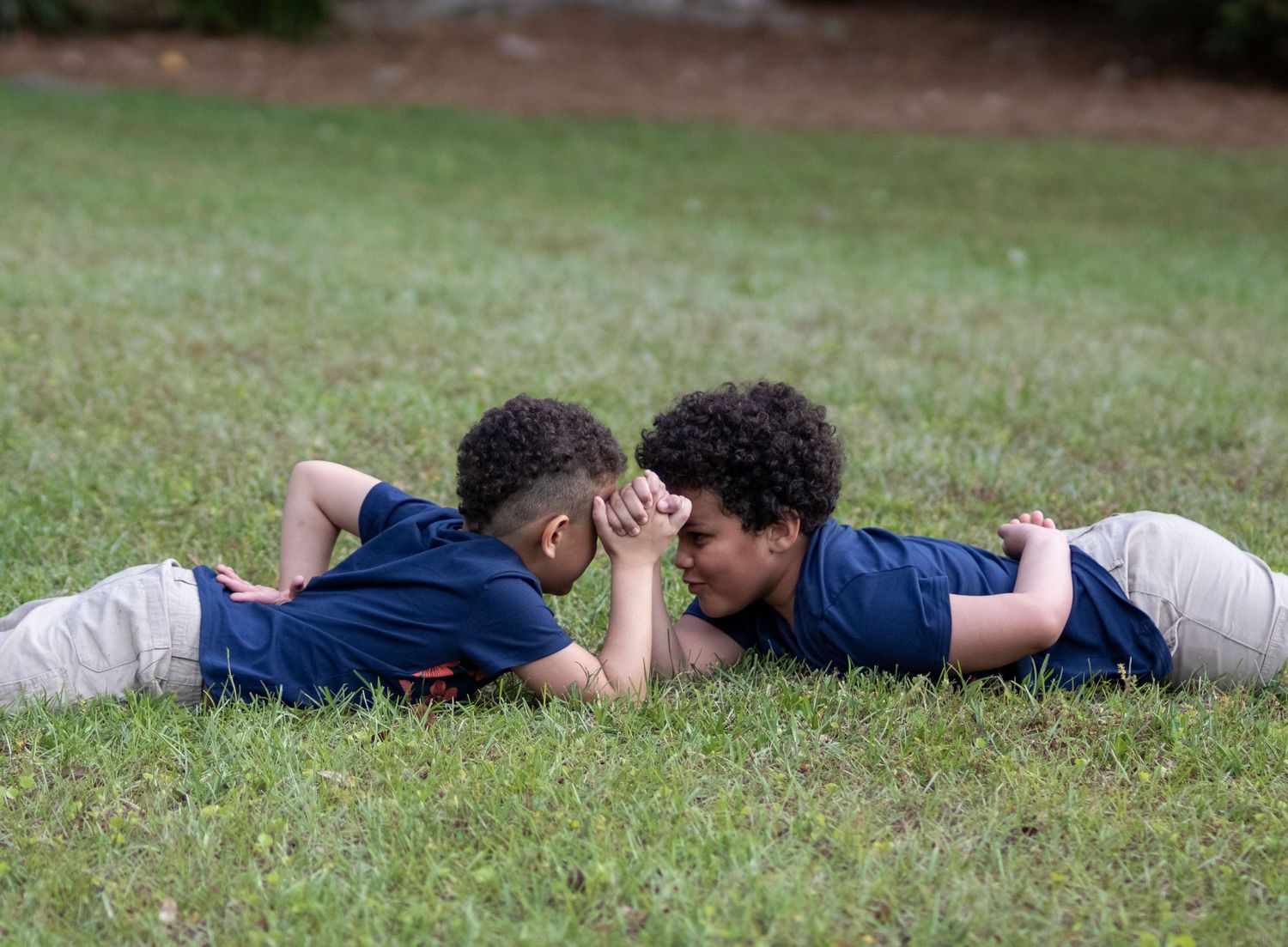 Two young boys lying on green grass face to face, holding hands in a heartwarming outdoor moment.