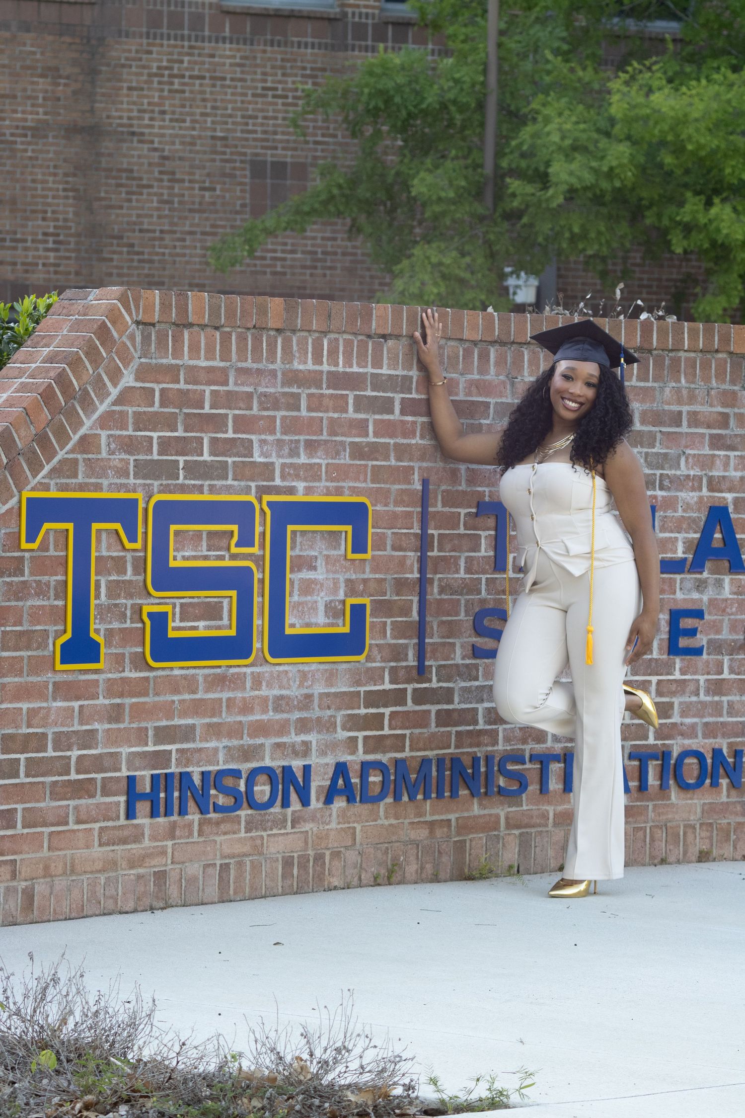 A proud graduate poses in a white outfit and cap beside the TSC Talladega State College Hinson Administration building sign.