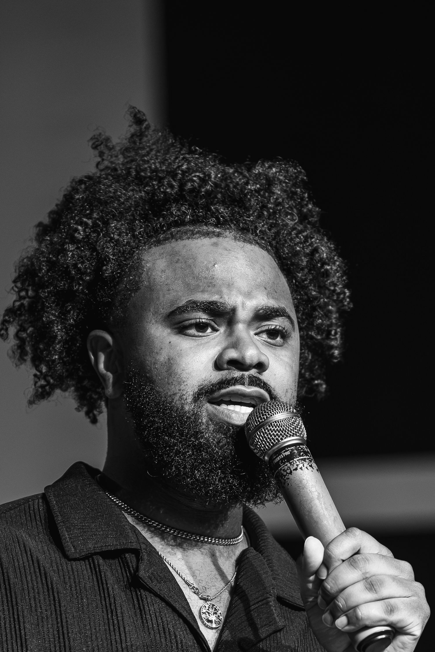 Black and white portrait of a young man with curly hair and beard speaking into a microphone.
