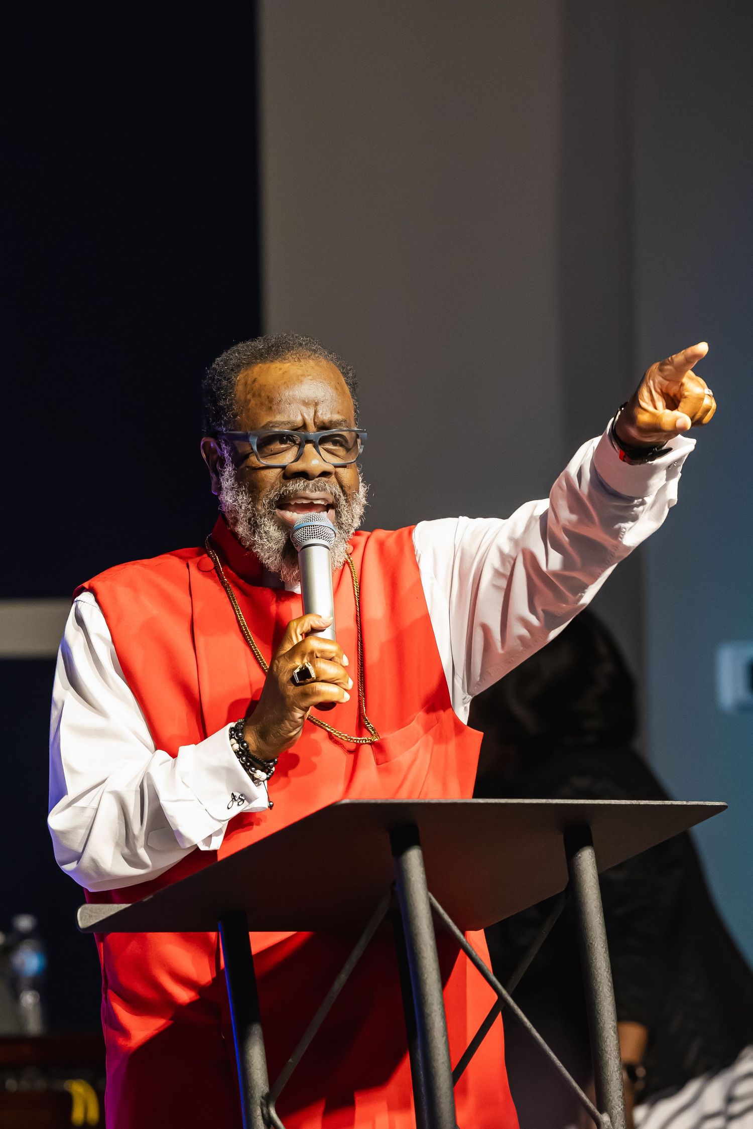 Elderly Black preacher in red vestments speaks passionately at podium, pointing finger while holding microphone.