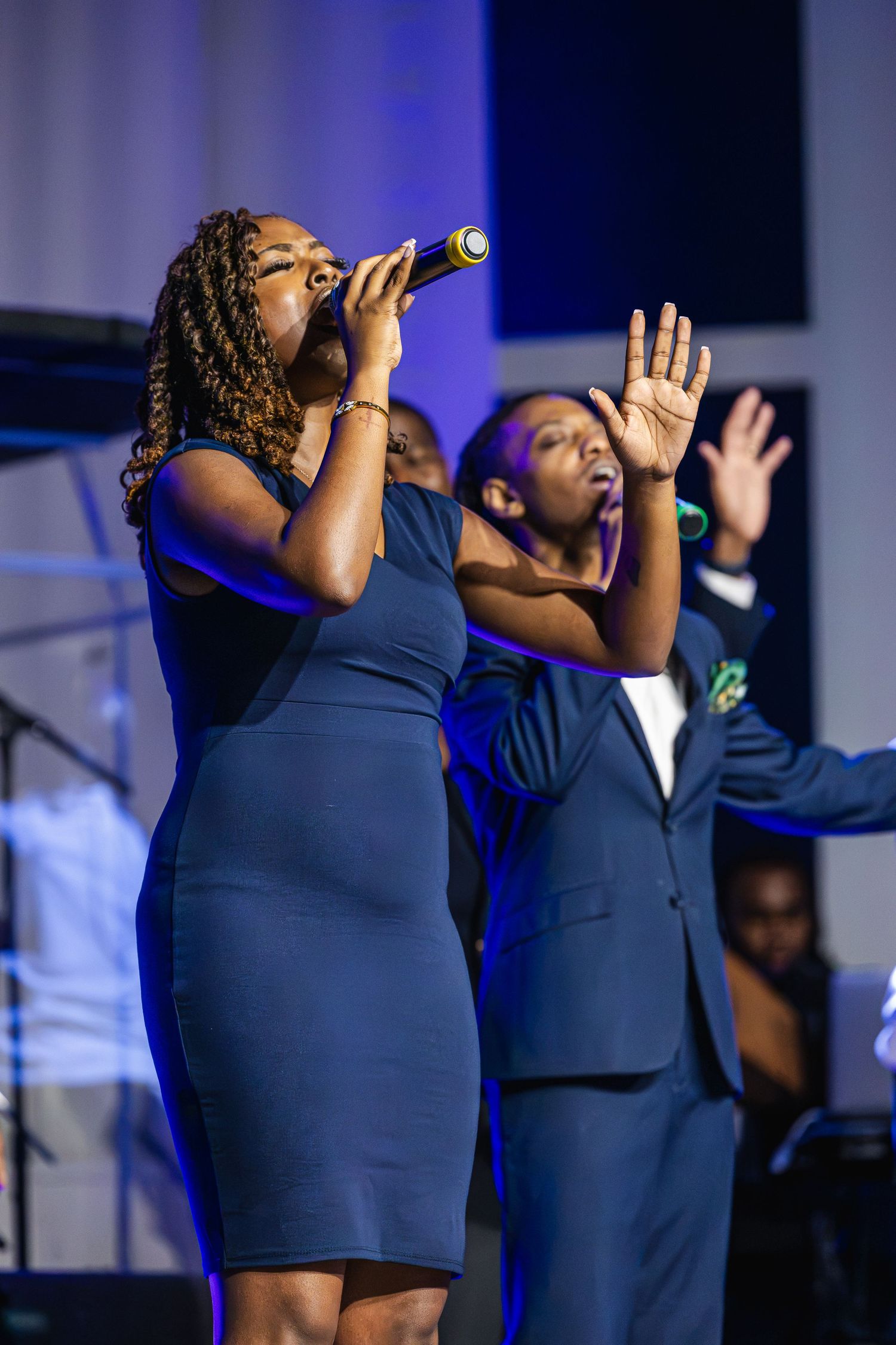Gospel choir performers in navy blue attire singing passionately on stage with dramatic blue lighting.