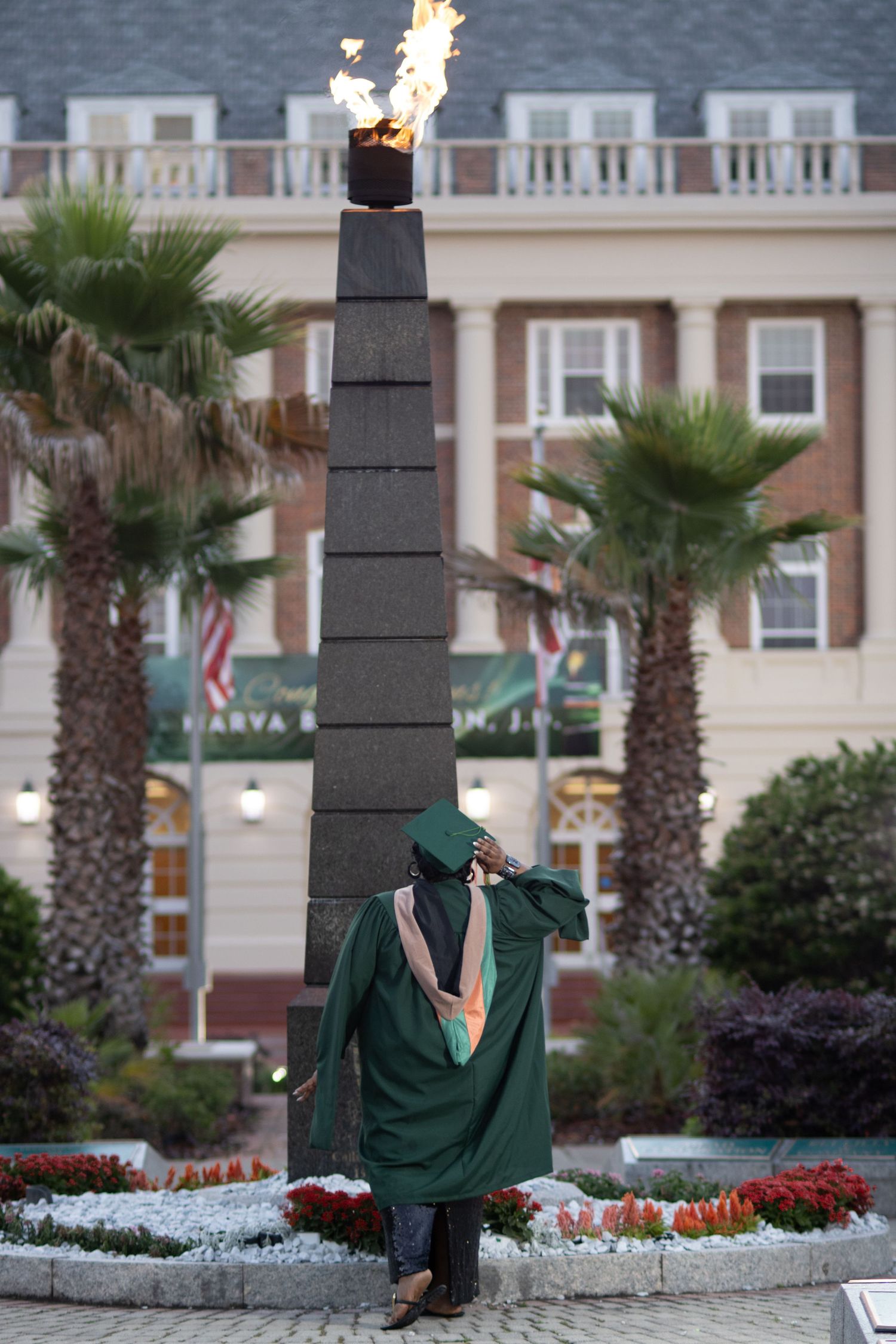 Graduate in green cap and gown stands before a tall stone torch monument with flame, flanked by palm trees.