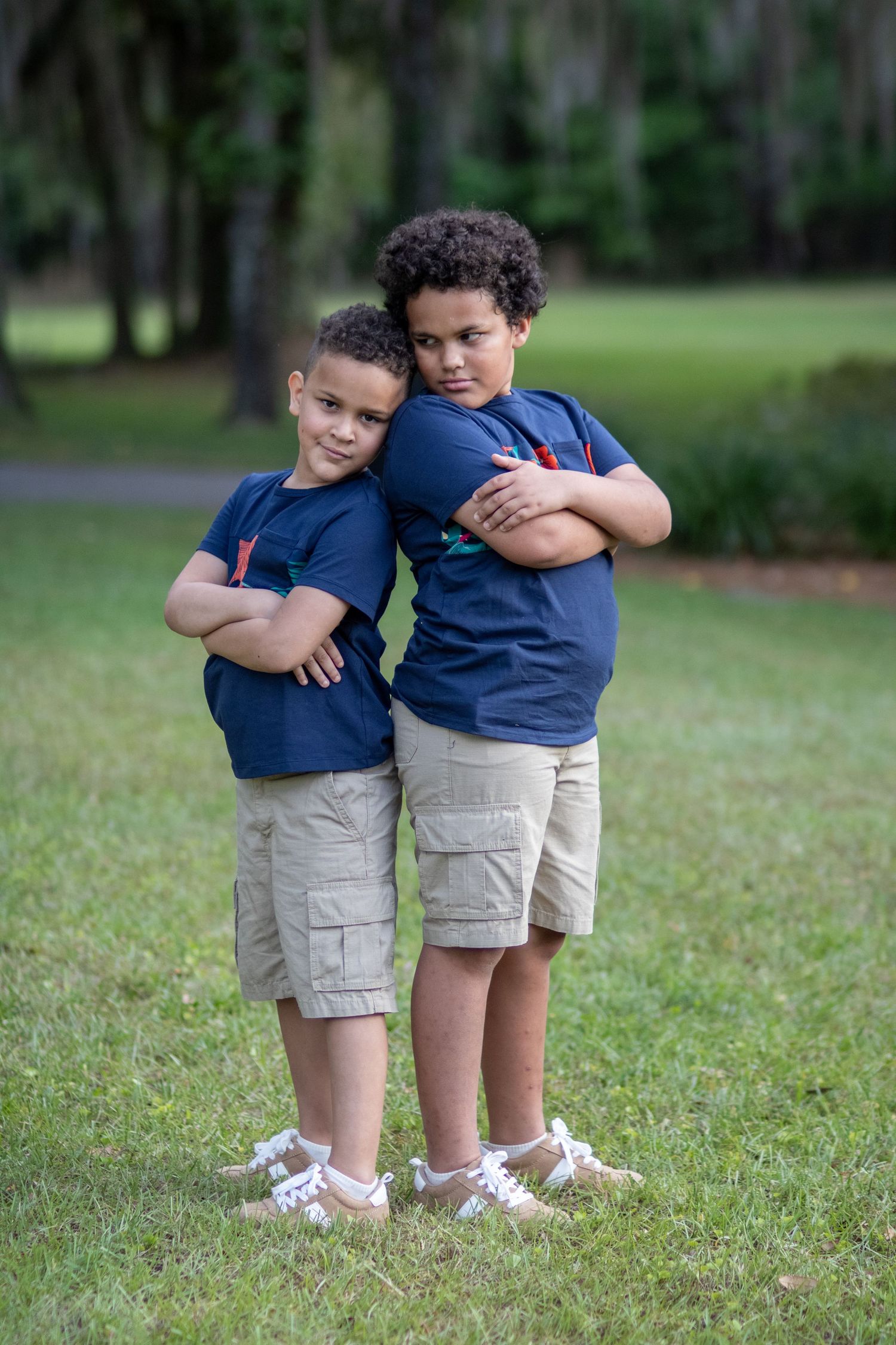 Two young boys in matching navy shirts and khaki shorts stand back-to-back with arms crossed in a park.