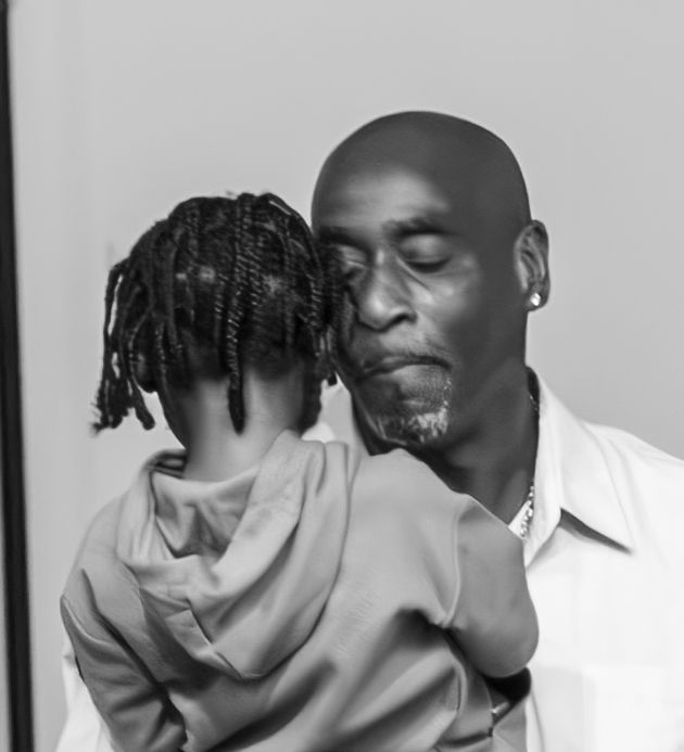 Black and white photo of a bald man tenderly kissing a child with braids wearing a hoodie.