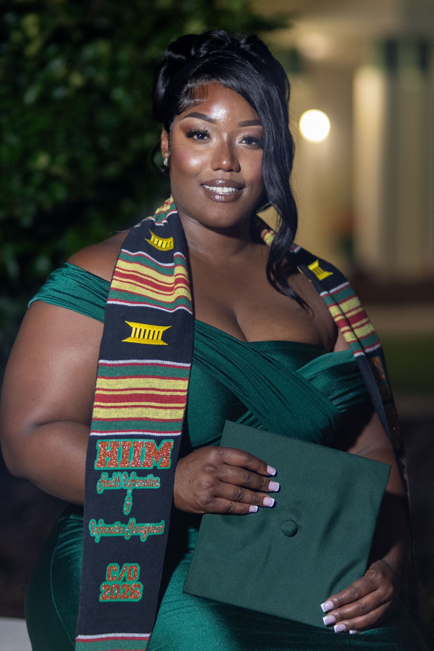 A young woman in a green off-shoulder dress holds a graduation cap, wearing a kente cloth stole at a nighttime ceremony.