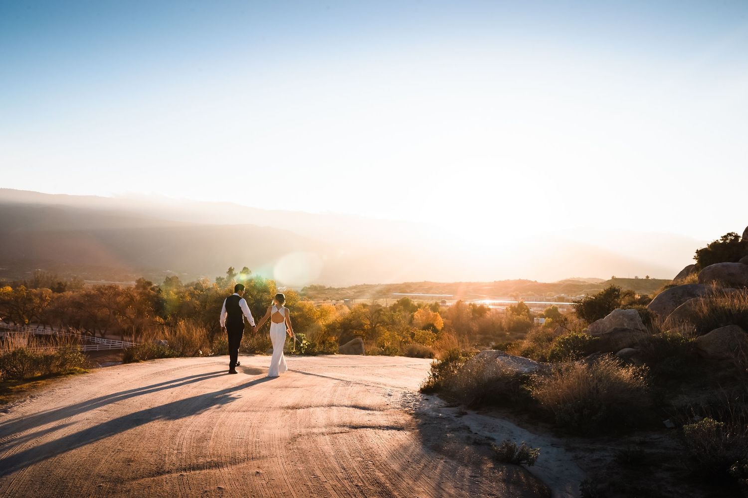 A couple walks on a winding road during sunset with golden mountains and sky in the background.