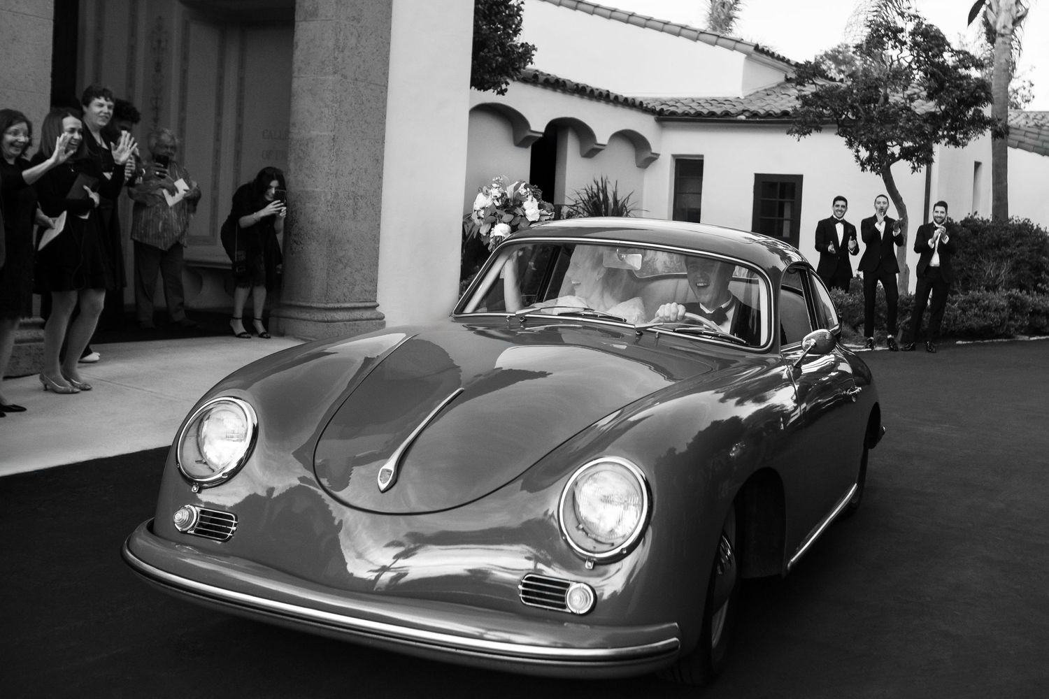 Classic Porsche 356 in black and white parked outside a vintage building with onlookers.