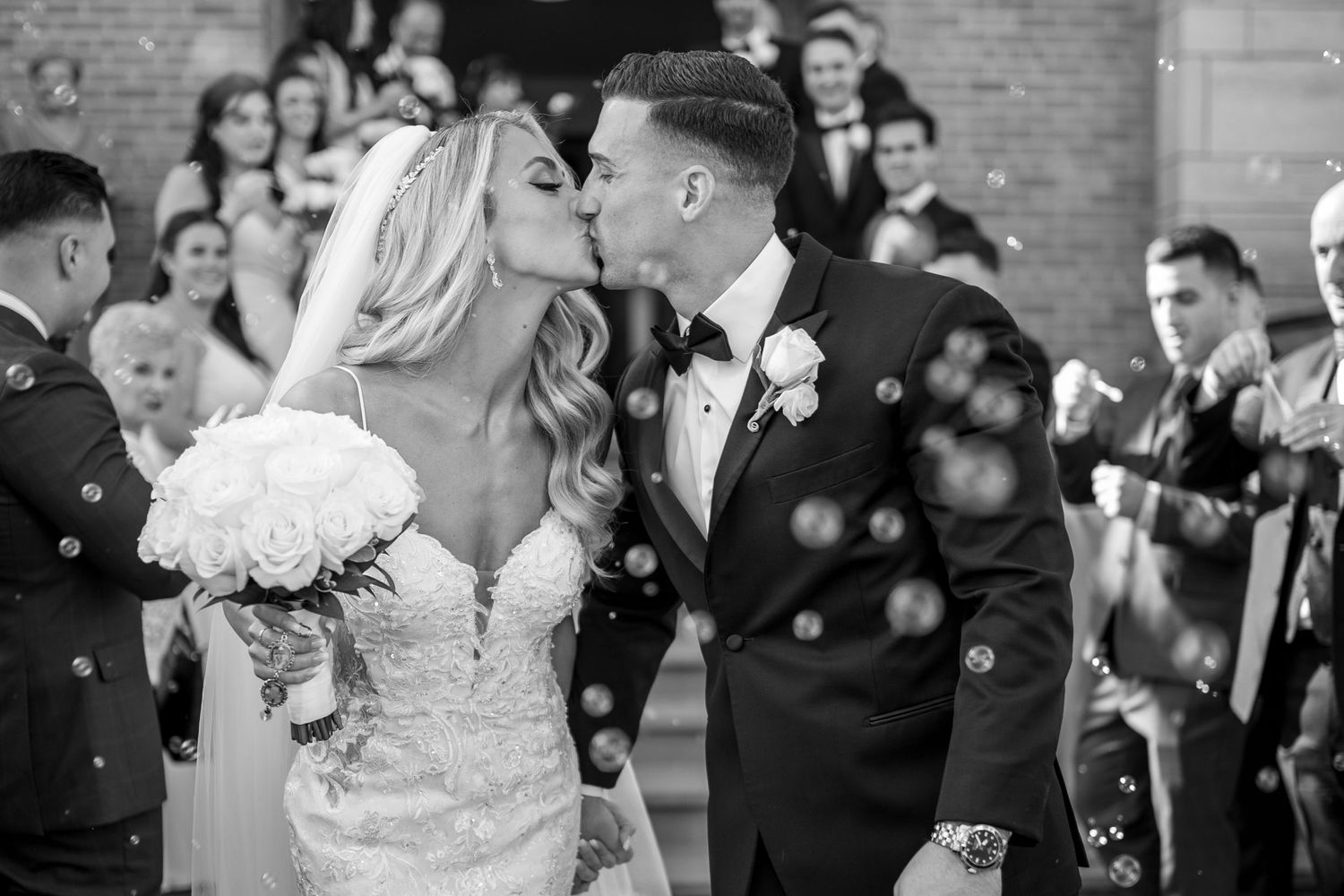 Wedding couple shares romantic kiss amid floating bubbles and guests.