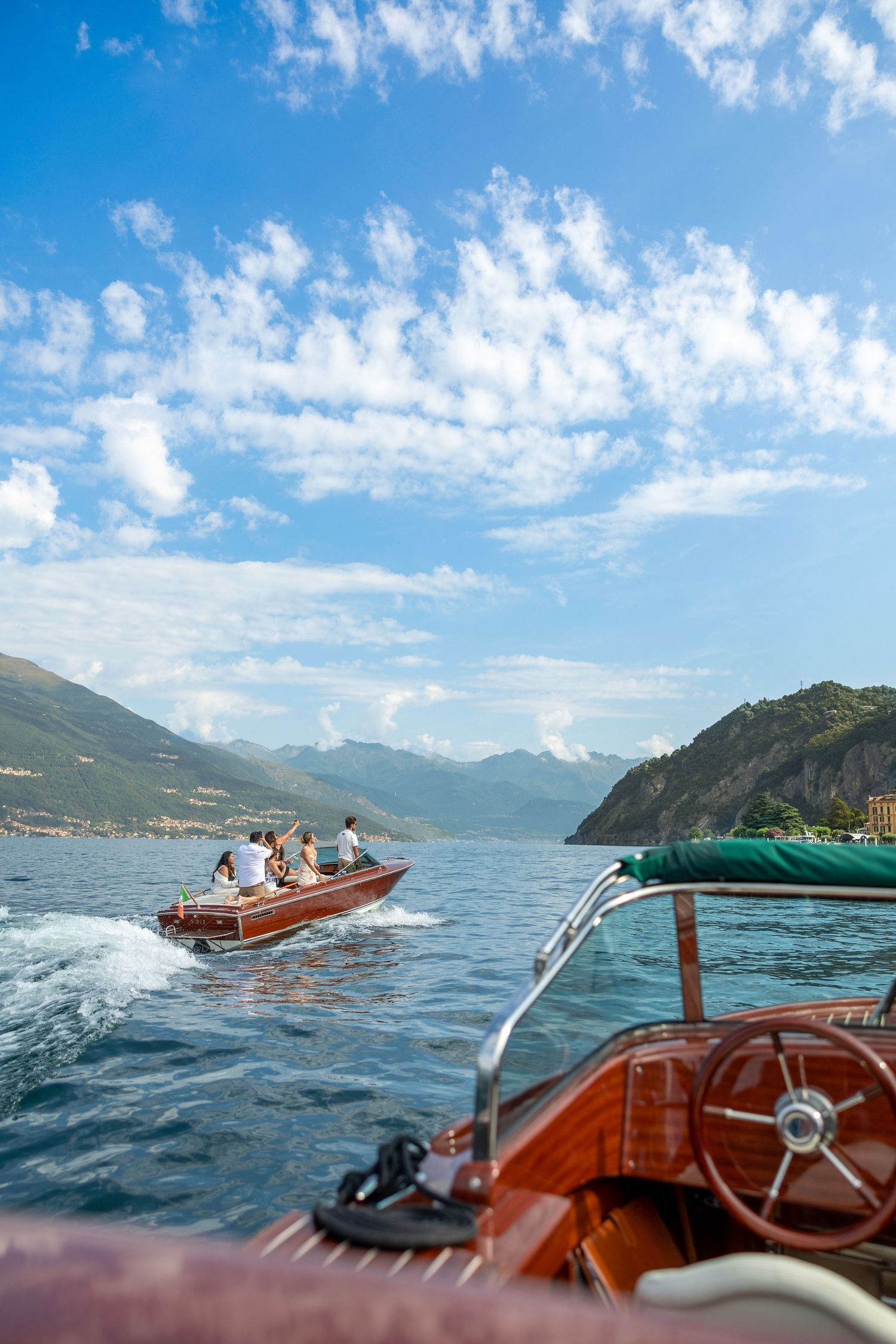 Wooden boats cruising on a scenic mountain lake under blue skies with puffy clouds in Lake Como, Italy.
