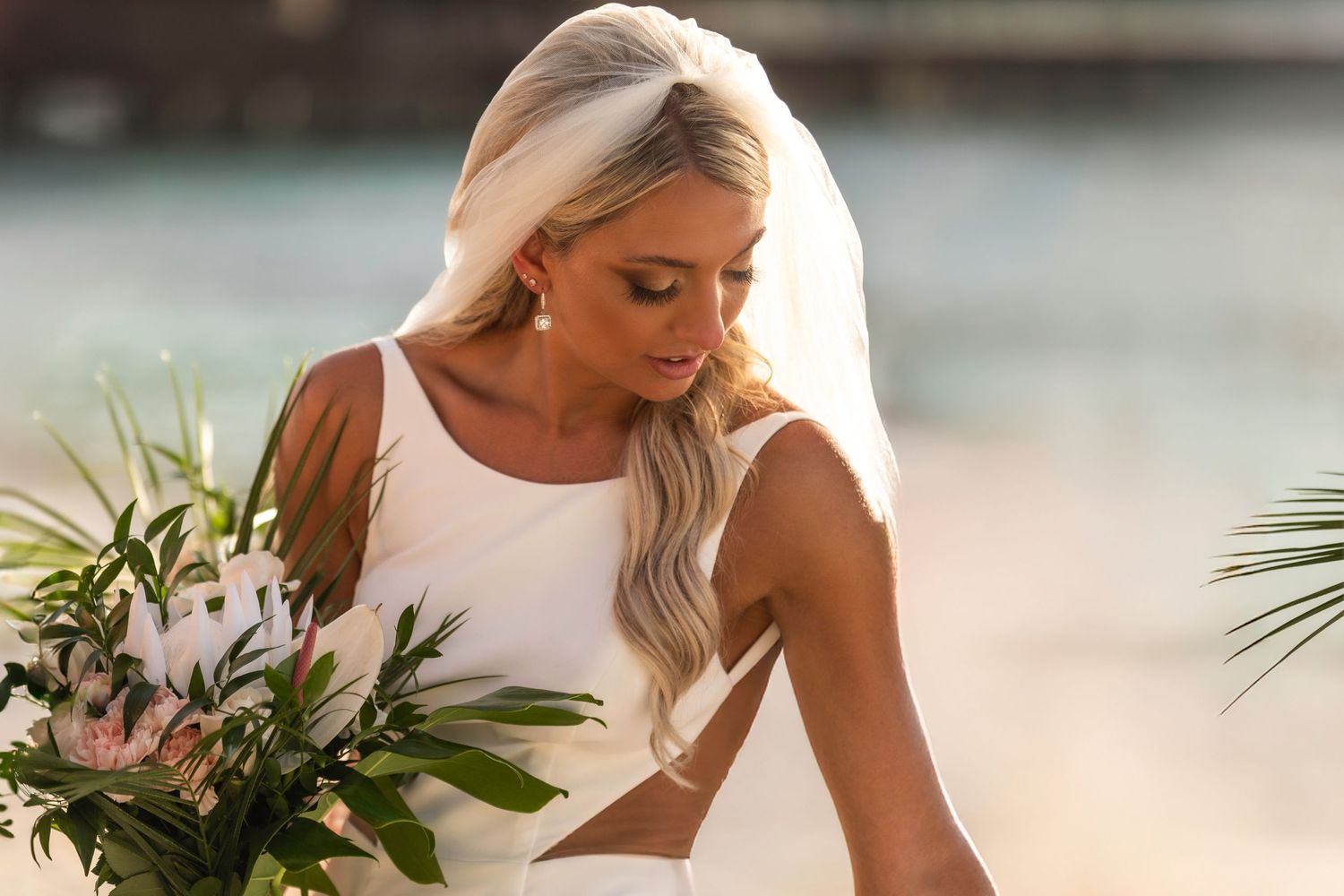 Bride in white dress holds tropical bouquet against sunset backdrop with palm fronds framing the scene.