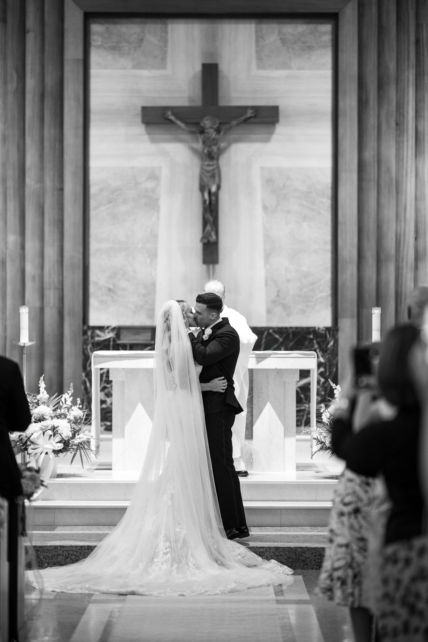 A couple shares their first kiss during a church wedding ceremony beneath a large crucifix.