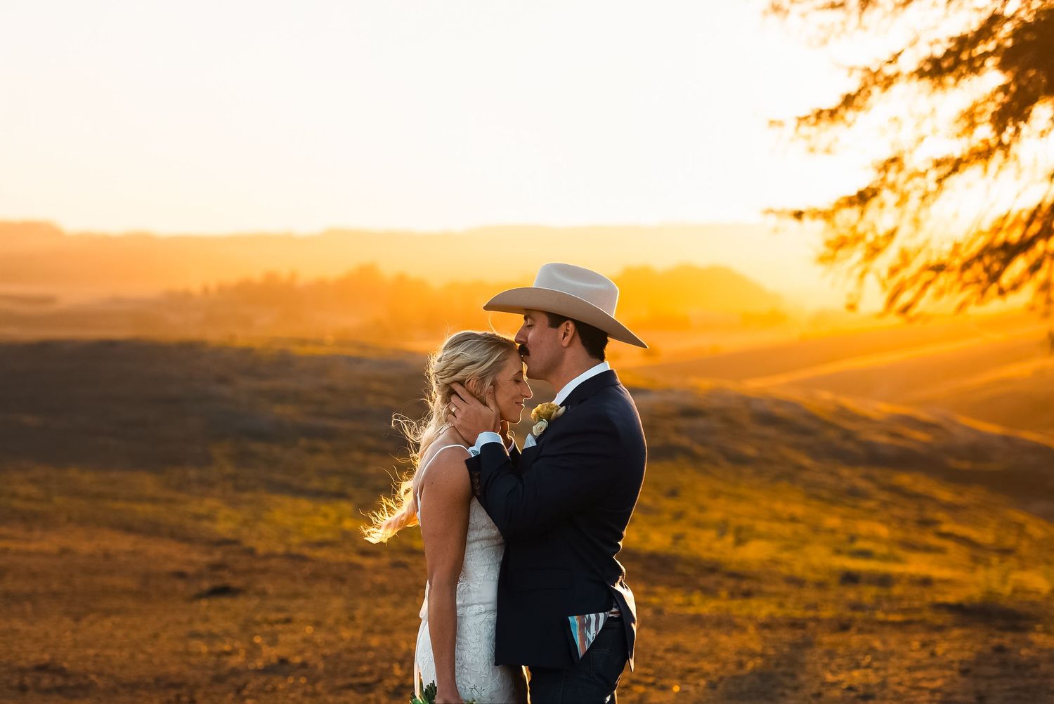 A couple in western attire embraces during a golden sunset over rolling hills in the countryside.