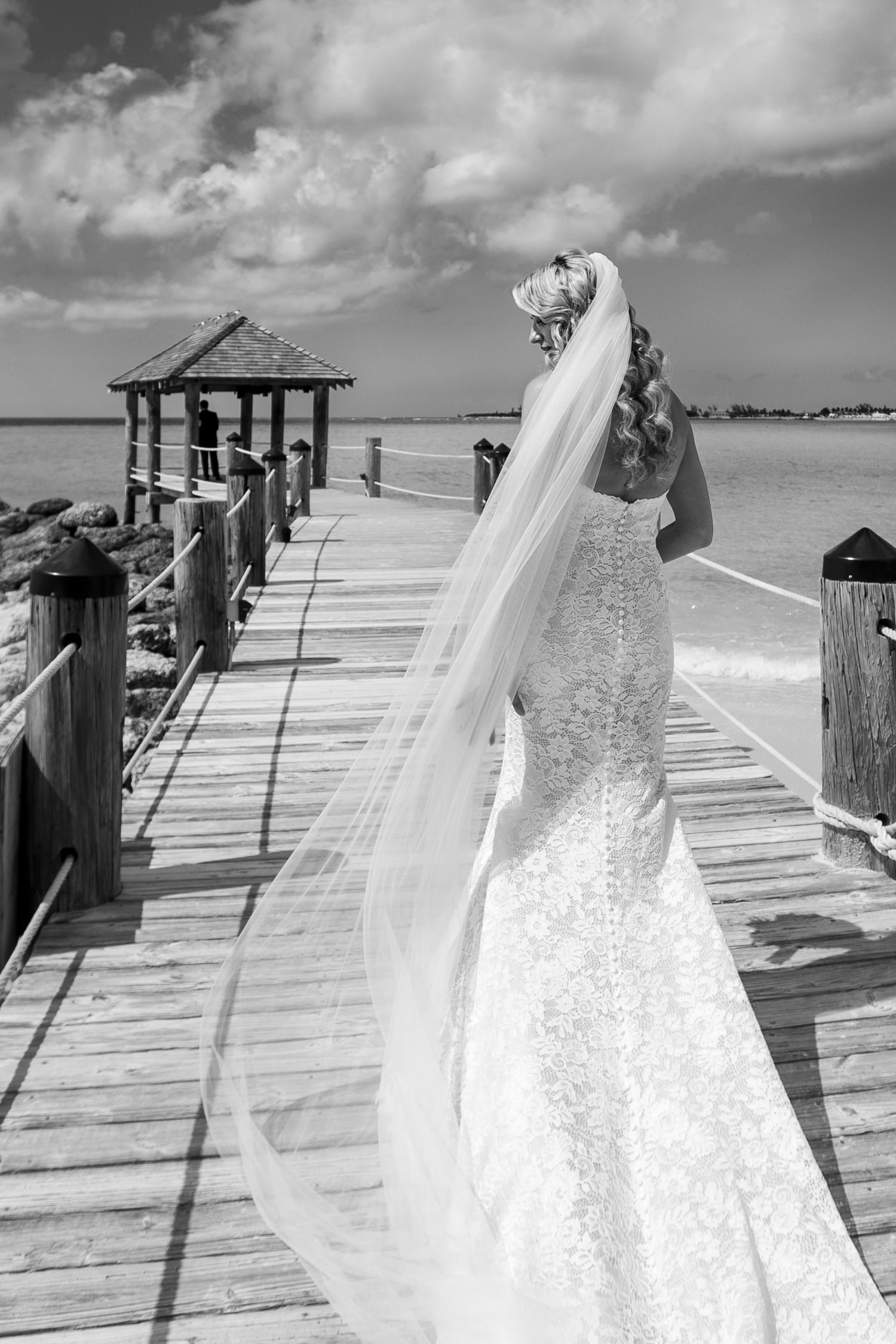 A stunning black and white photograph of a wedding dress and veil on a beachside boardwalk with a gazebo in the distance.