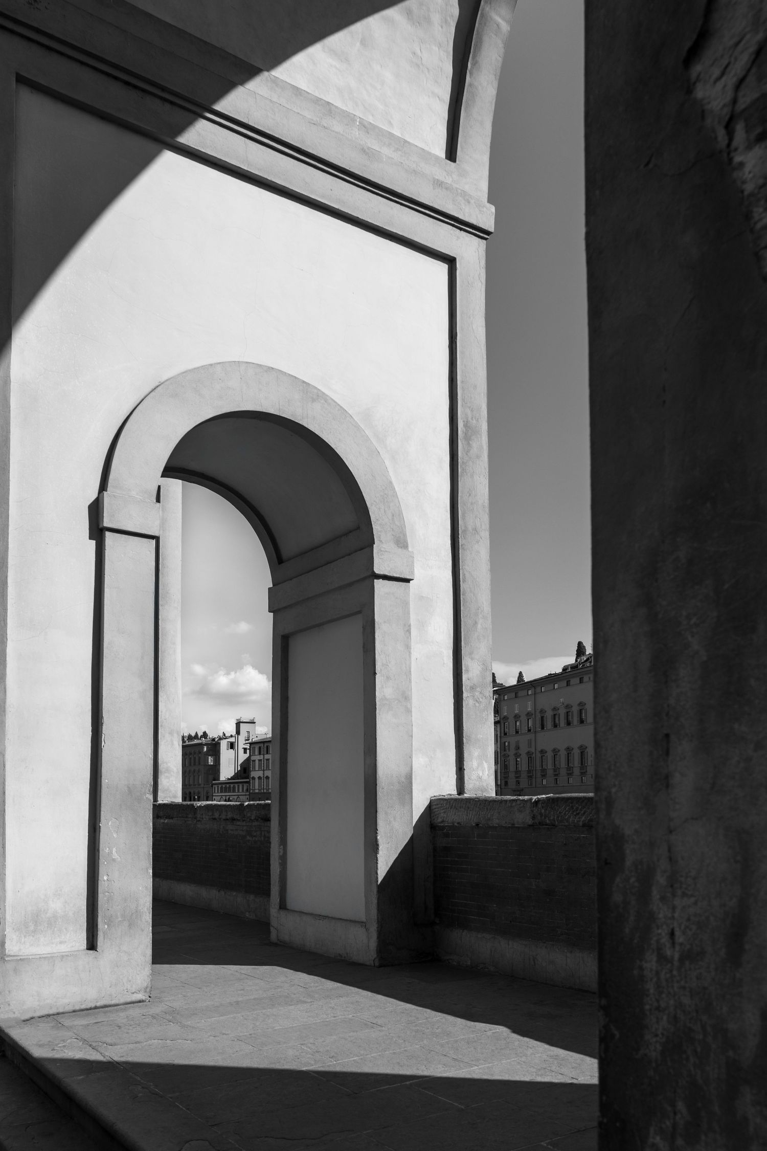 Sunlight creates dramatic shadows through a white stone archway overlooking a city.