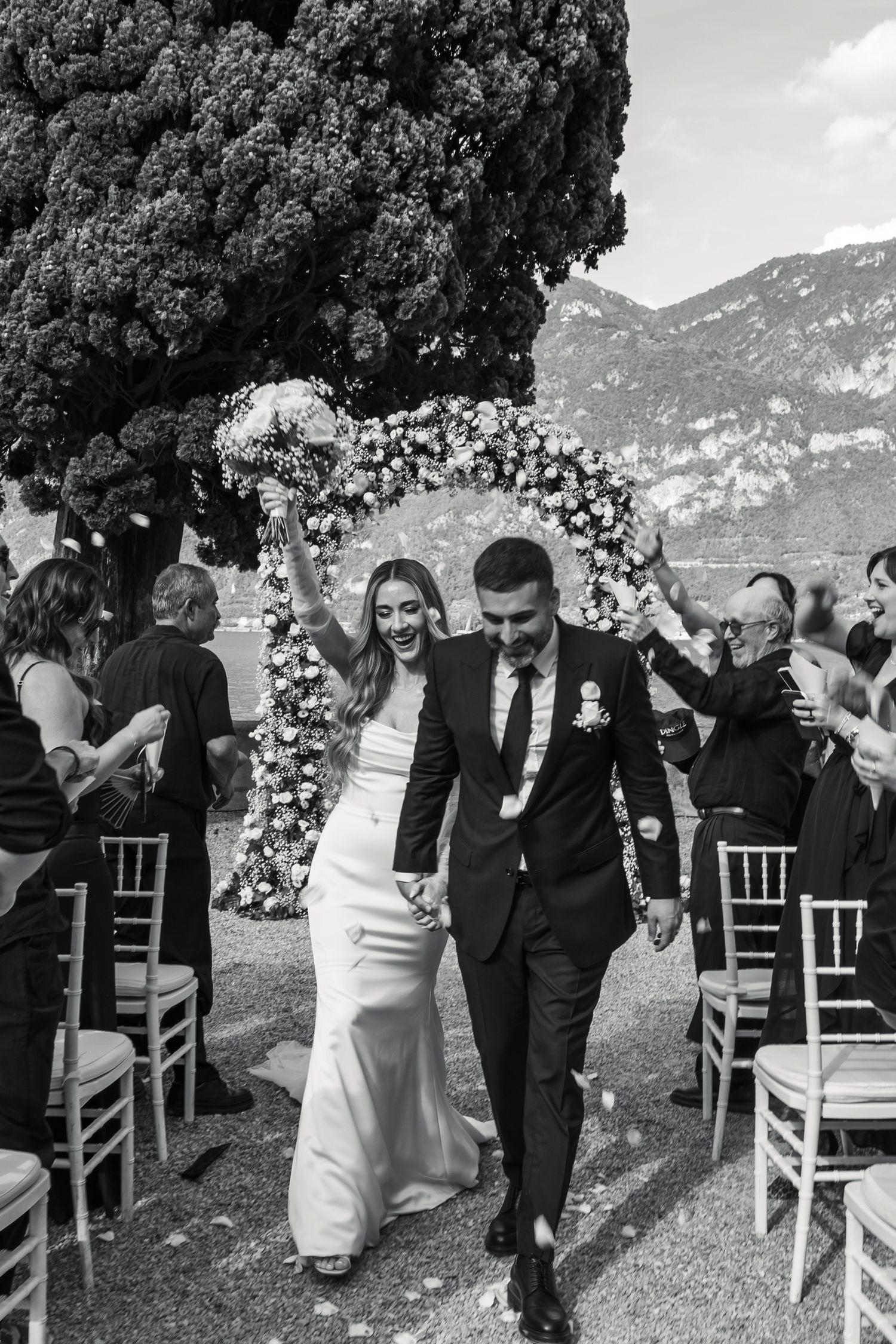Black and white wedding photo of a couple walking down the aisle under a floral arch with Lake Como and mountains in the background.