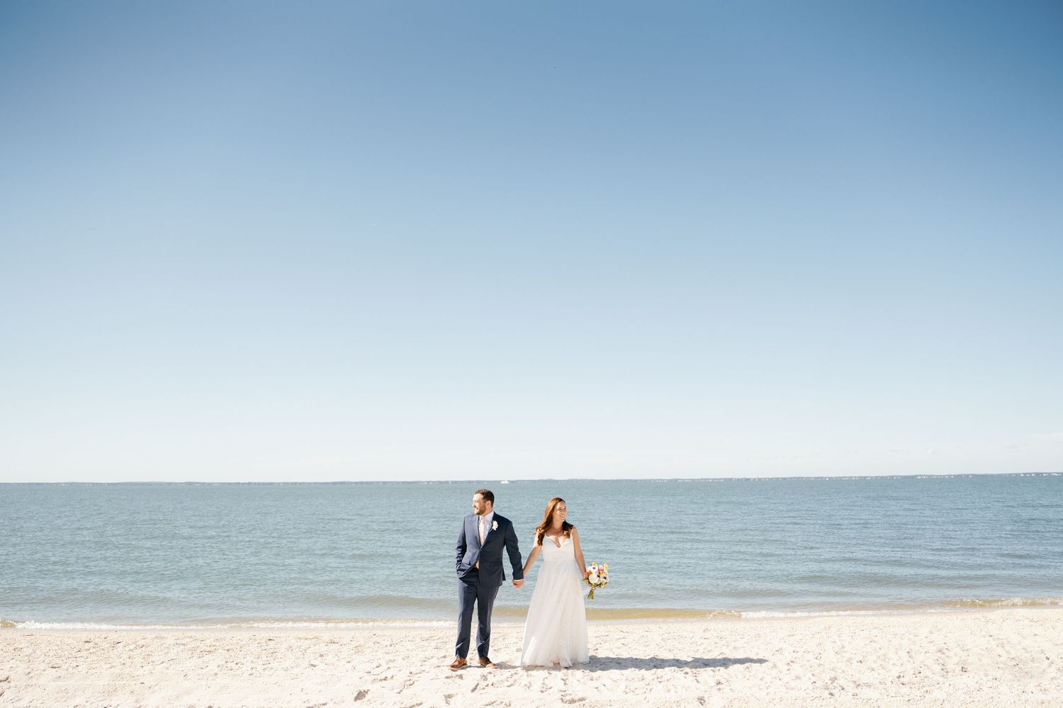 A couple walks along a scenic beach shoreline with calm waters and blue skies in the background.