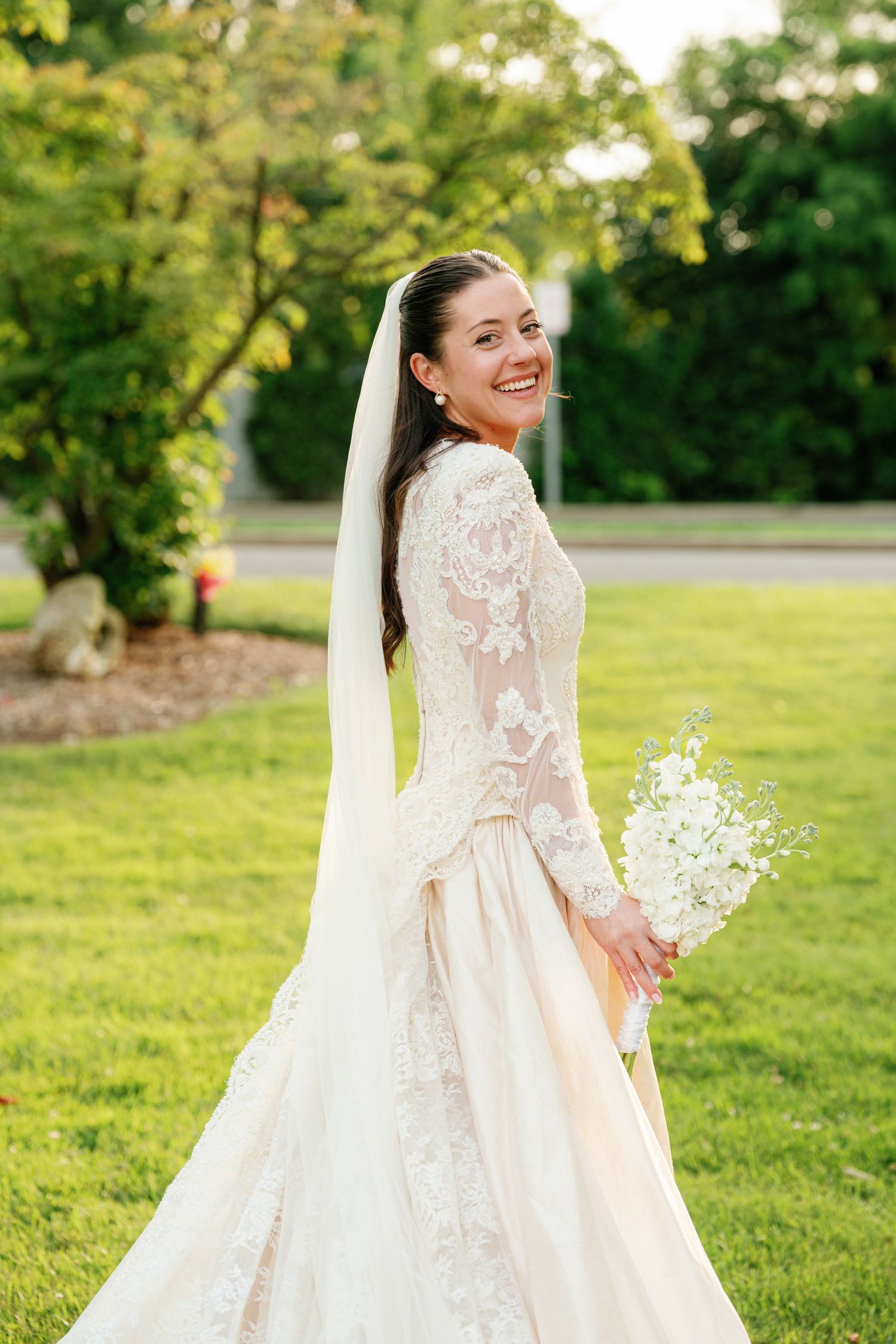 A bride in an elegant cream lace gown poses joyfully in a sunlit garden setting with flowing veil.