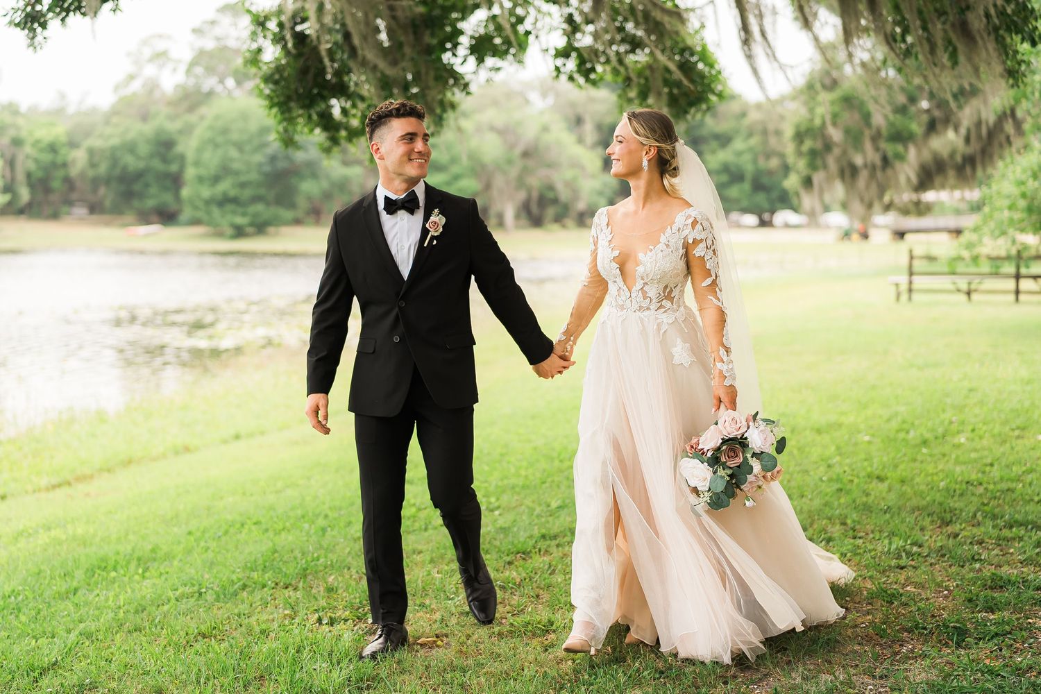 A couple in formal wedding attire walks hand in hand on grass near a waterfront during an outdoor celebration.