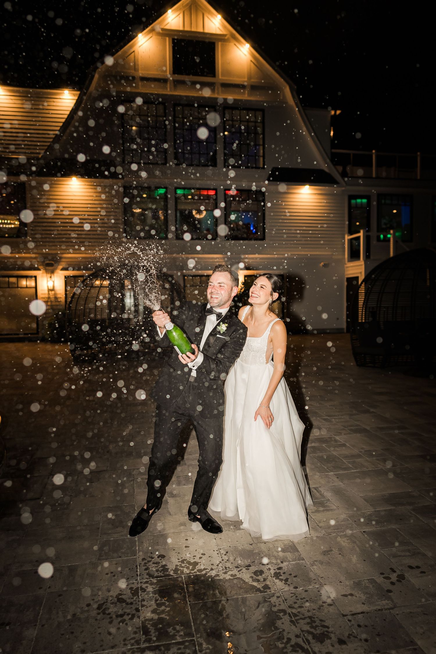 Couple sprays champagne in snowy nighttime celebration outside rustic venue.