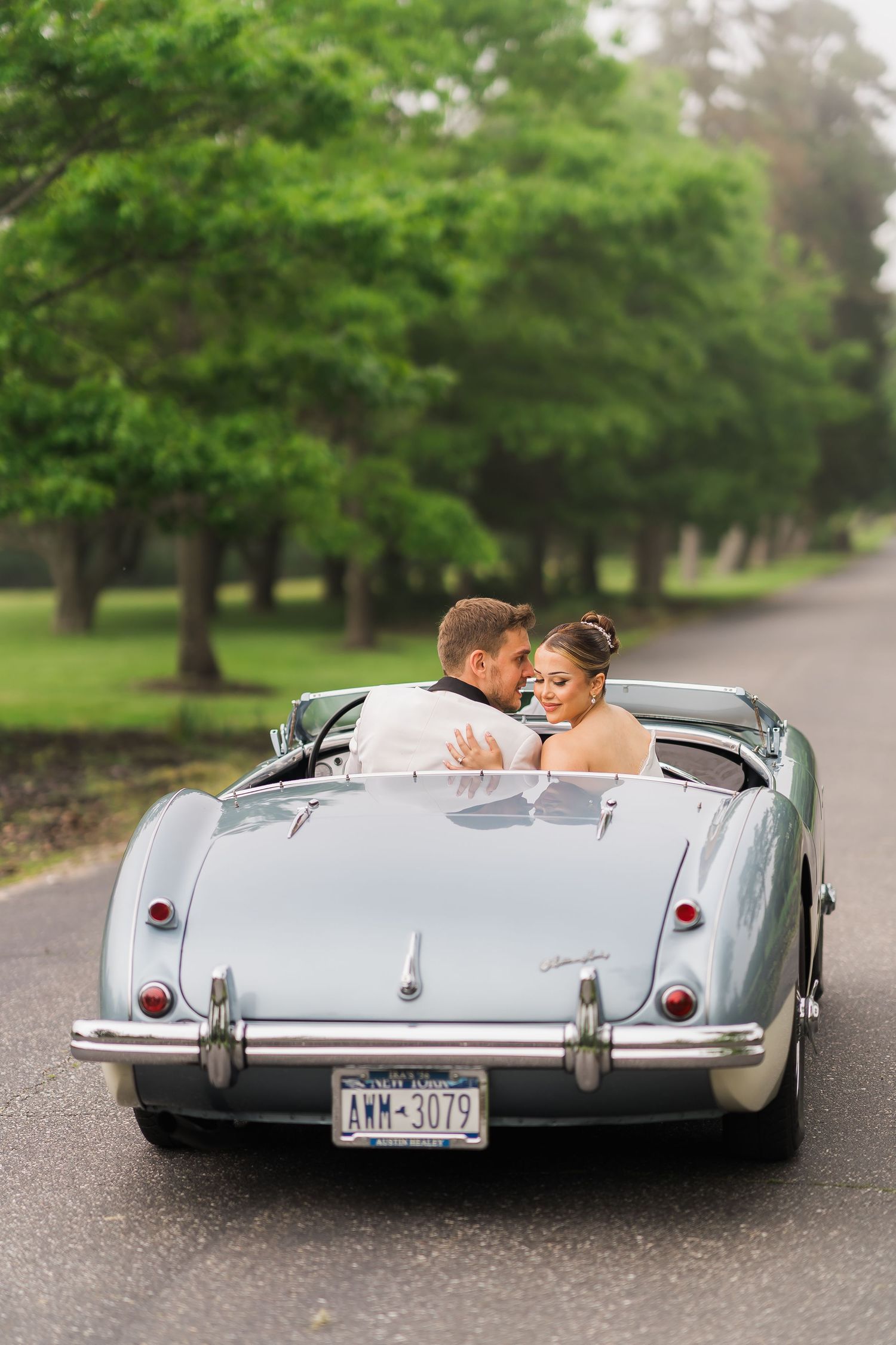 A couple embraces in a classic silver convertible car on a tree-lined road.