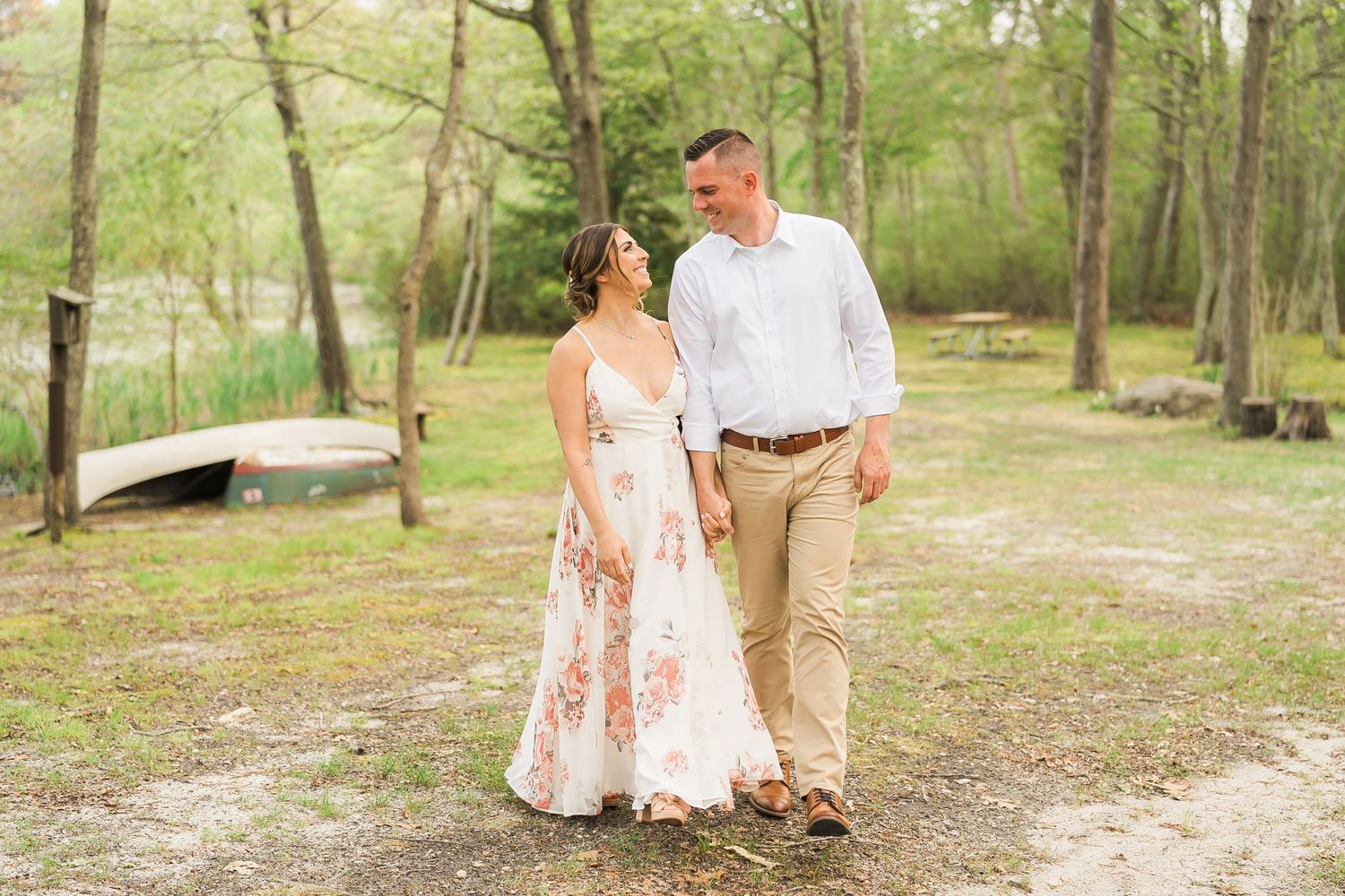 A couple walks together on a wooded path near a lake during an engagement photo session in springtime.