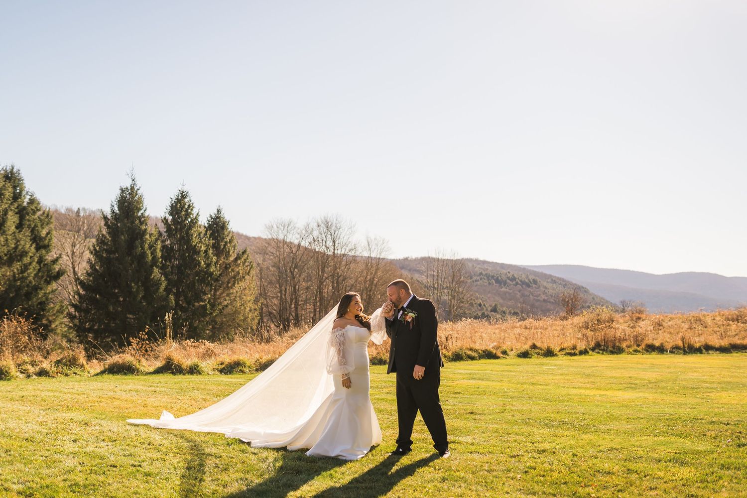 A wedding couple walks across a scenic grassy meadow with mountains and evergreen trees in the background.
