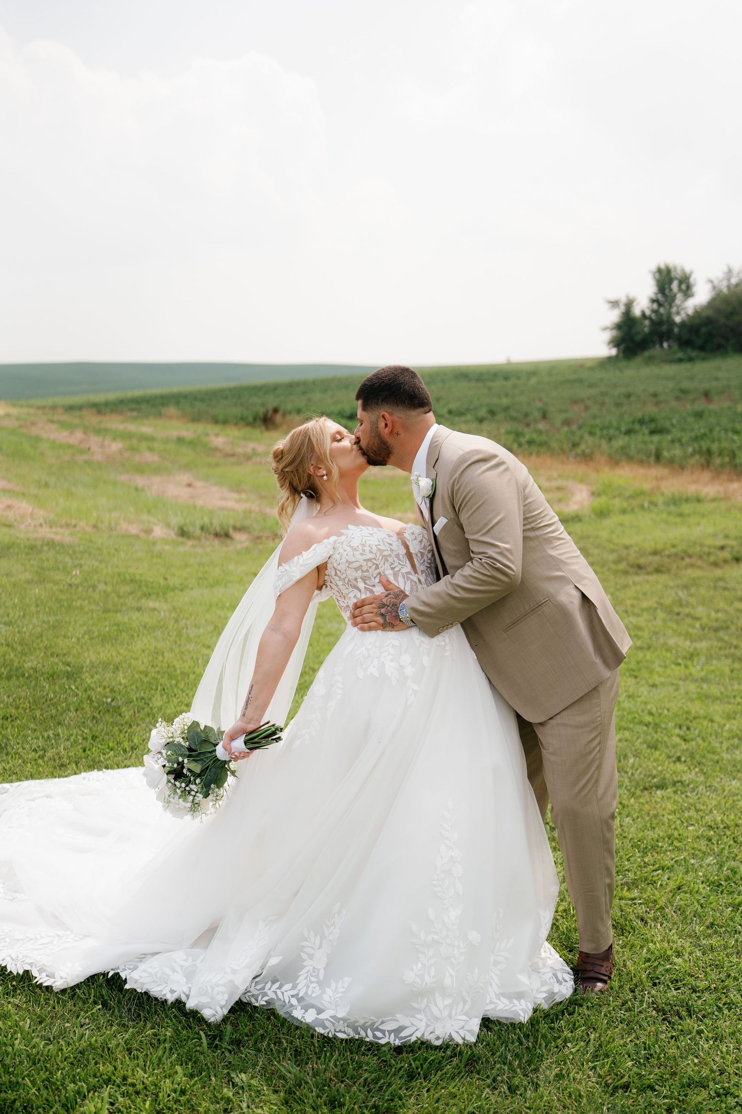 A couple in wedding attire share an intimate moment on a grassy hillside in a romantic outdoor setting.