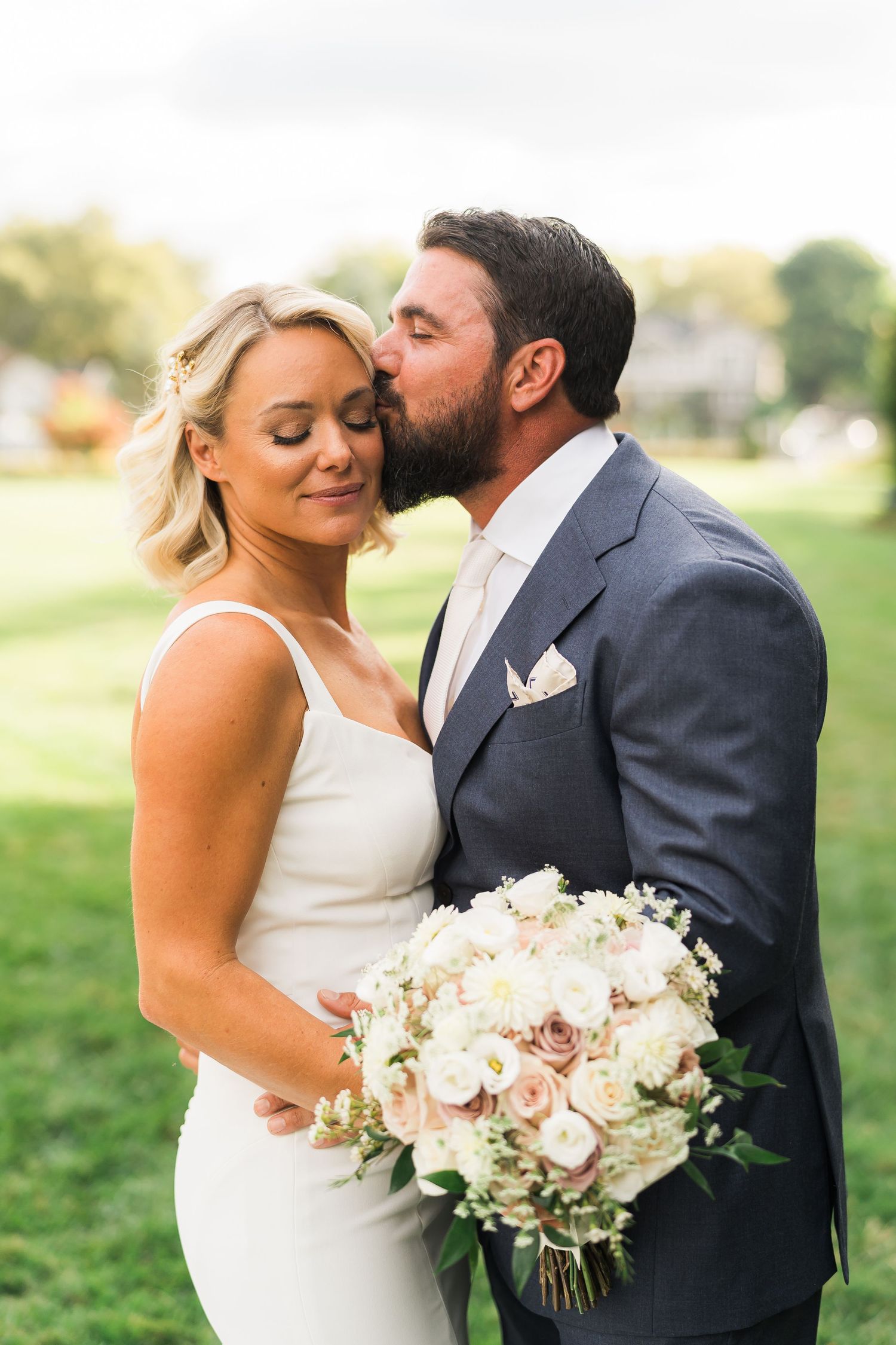 A romantic wedding photo of a couple with a blush pink and white rose bouquet in an outdoor garden setting.