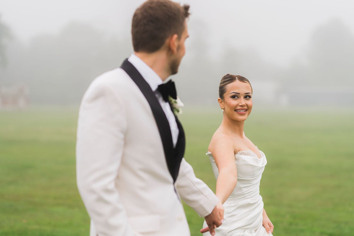 A couple in wedding attire stands together in a misty field on their special day.