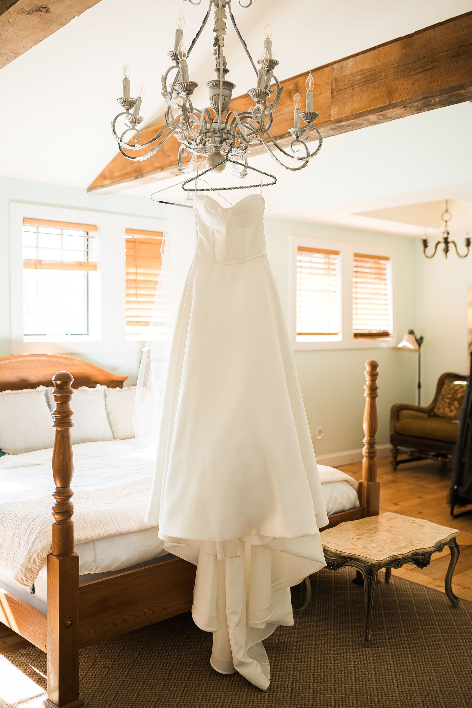 An elegant white wedding gown hangs from a vintage chandelier in a rustic room with wooden beams.