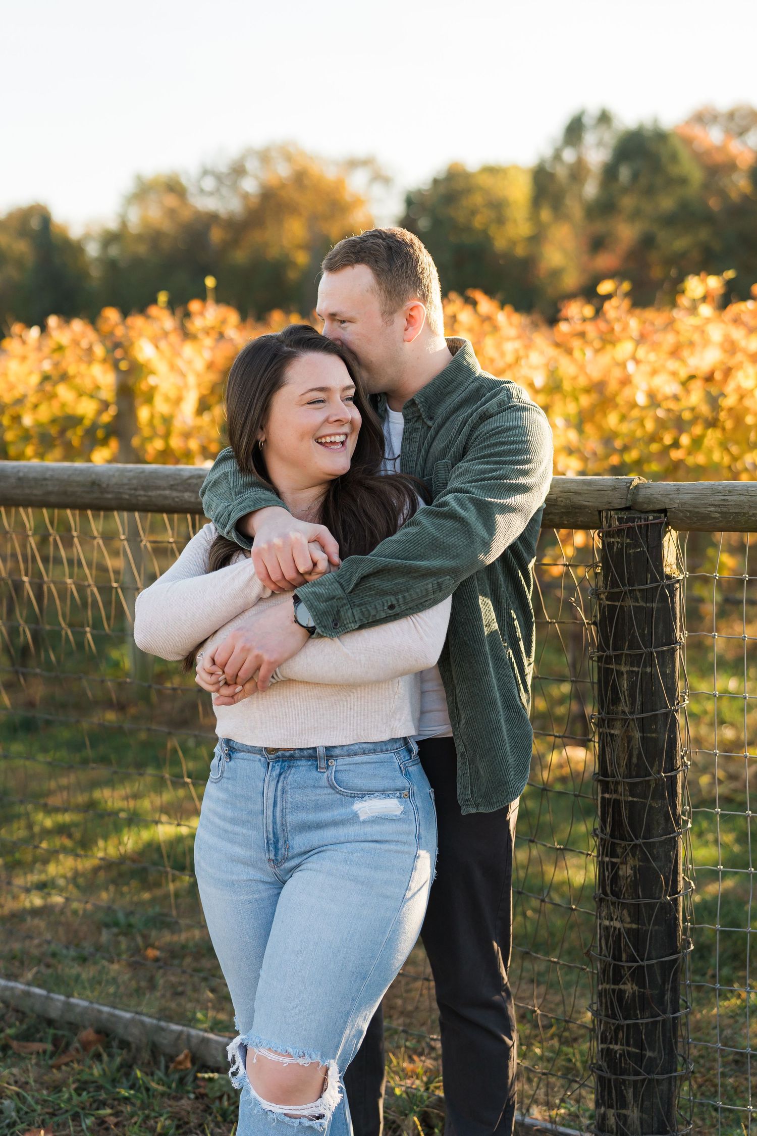 Happy couple shares a joyful embrace by a rustic fence with fall foliage in the background.